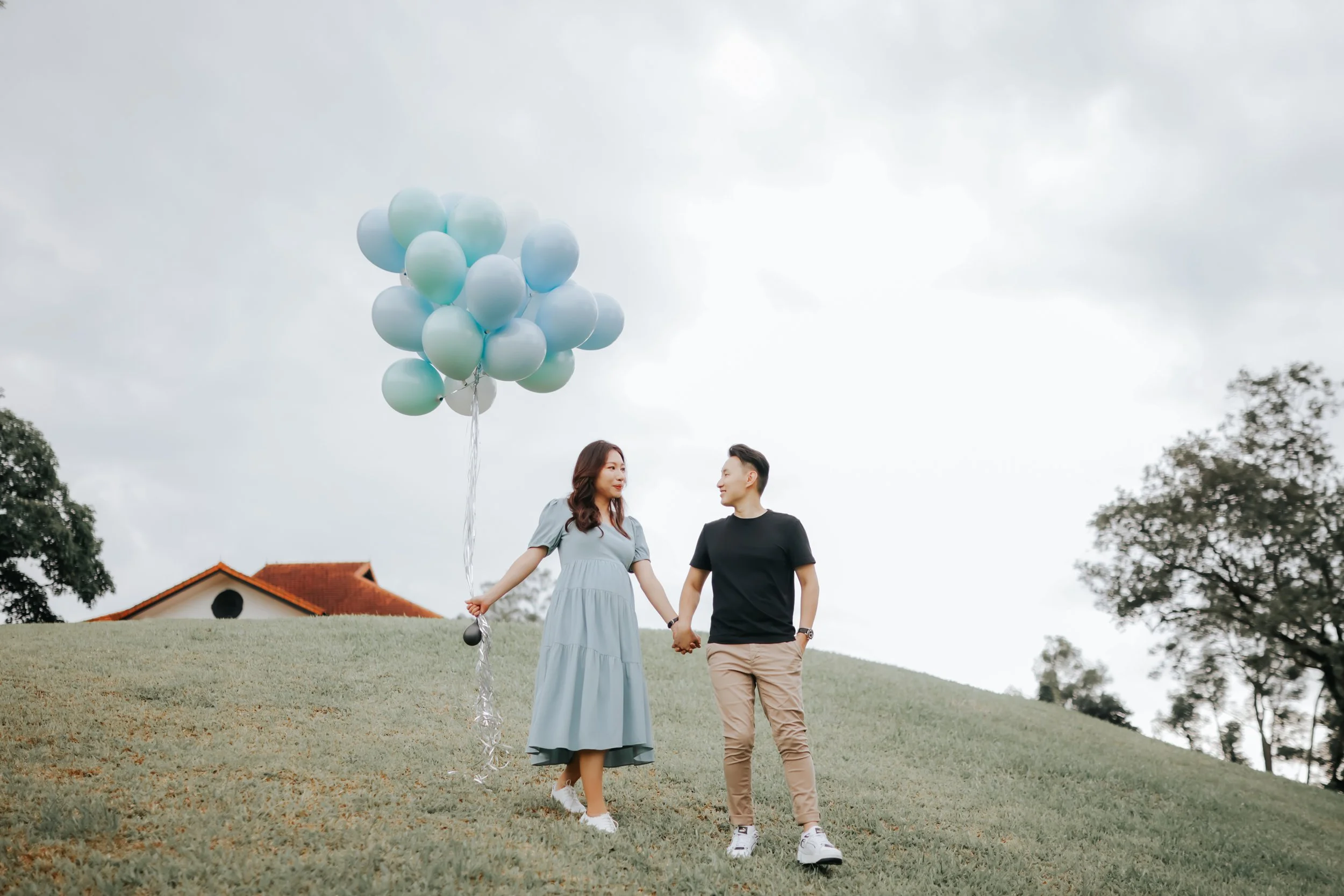 A couple on their maternity shoot captured walking down a hill at Gallop Extension Singapore while holding on to a bunch of balloons, captured by Breadandbutterworks in a soft and heartfelt style.