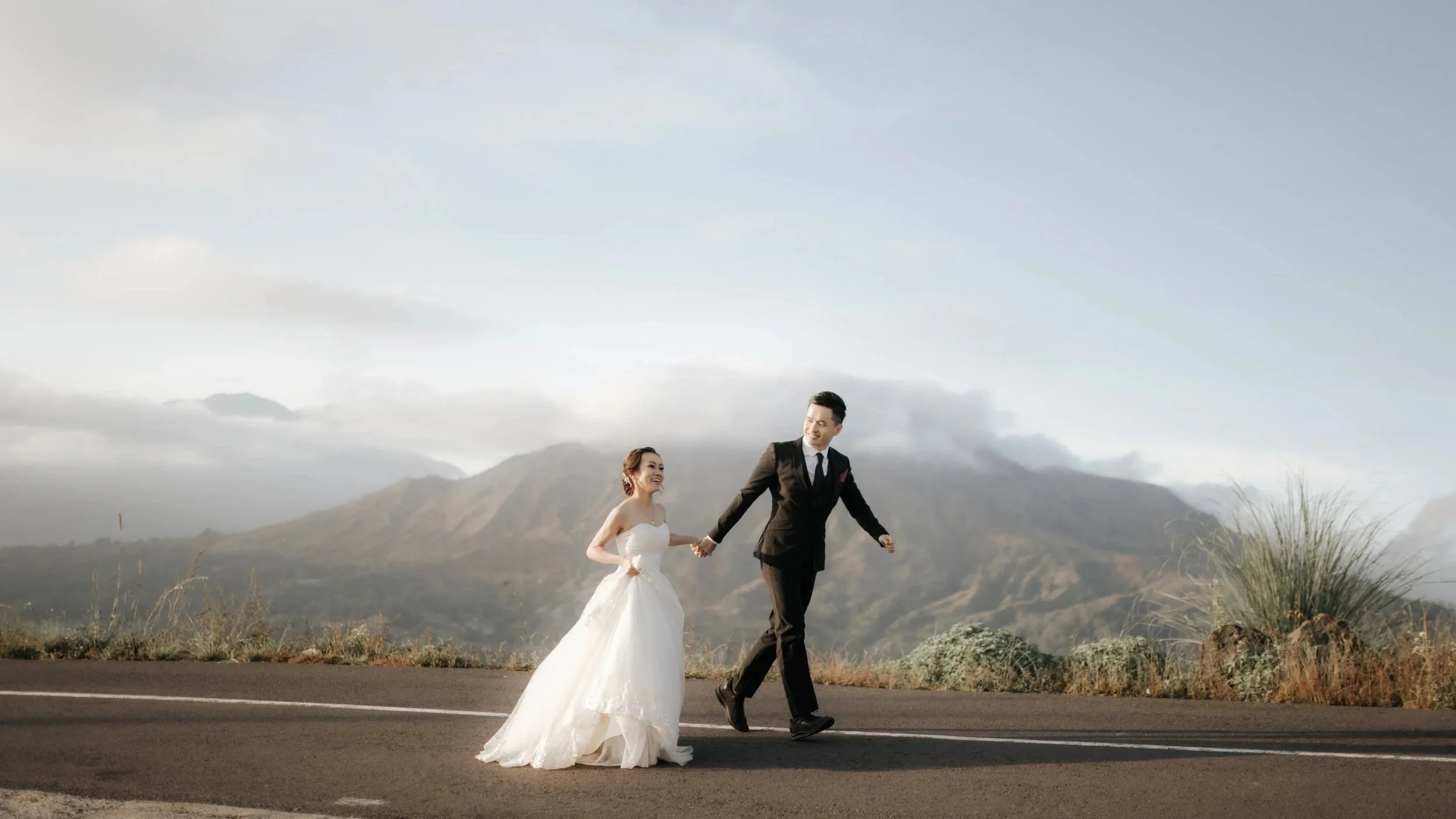 A bride and groom holding hands and walking along a road in a mountainous landscape in Bali during daytime. The bride is wearing a white dress and the groom is dressed in a black suit with a tie. They are smiling and looking at each other.