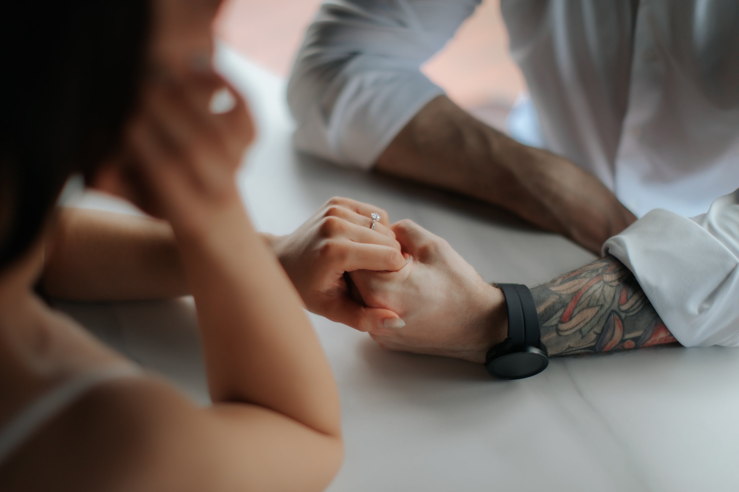 Cinematic light and airy proposal photoshoot. The photo features a close-up of the couple holding hands on a table, and the newly worn engagement ring glitters on the bride-to-be's finger. Captured by Arielle for Breadandbutterworks.
