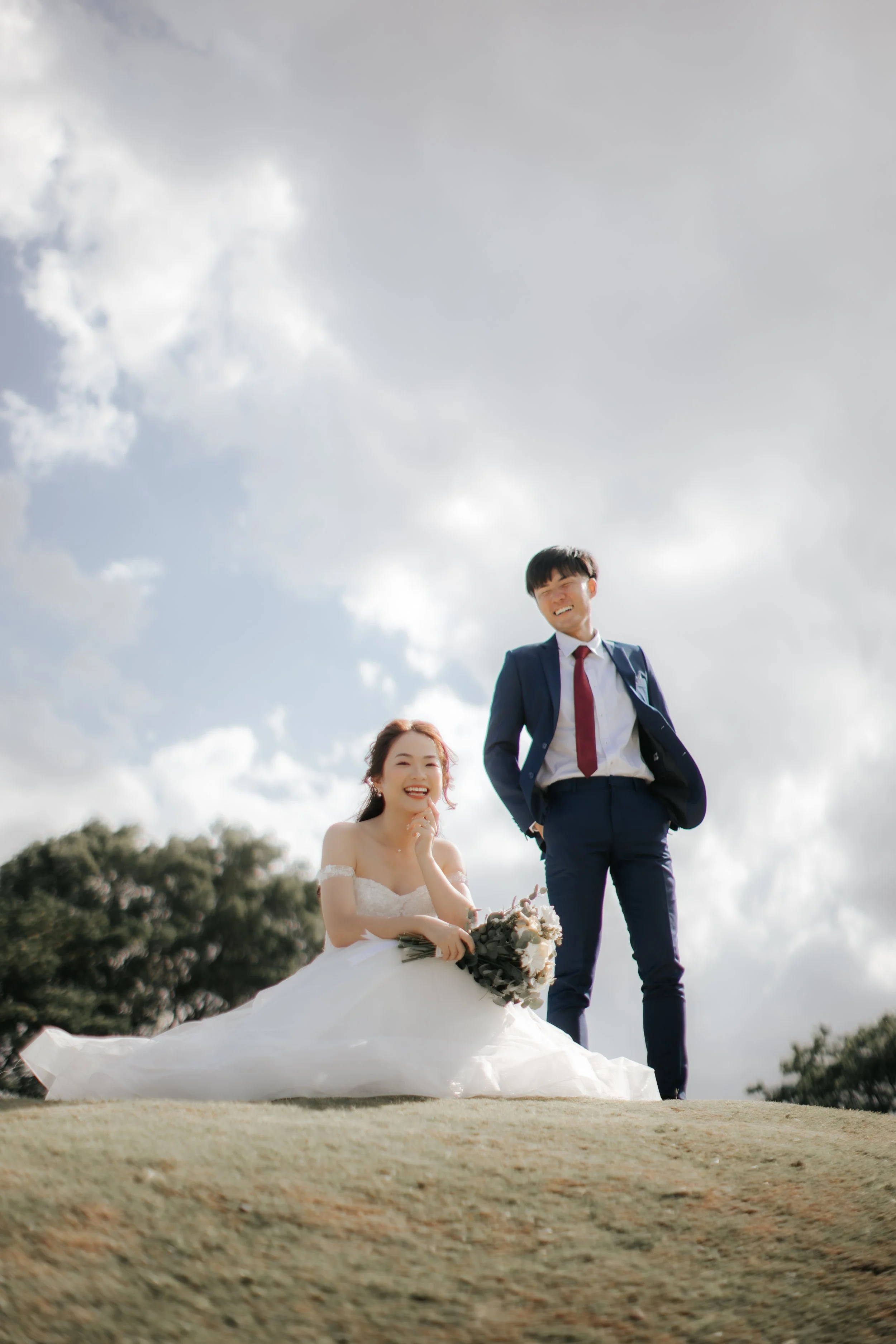 Light and airy pre-wedding photo of a bride and groom posing together under a dramatic sky in an outdoor Singapore setting.