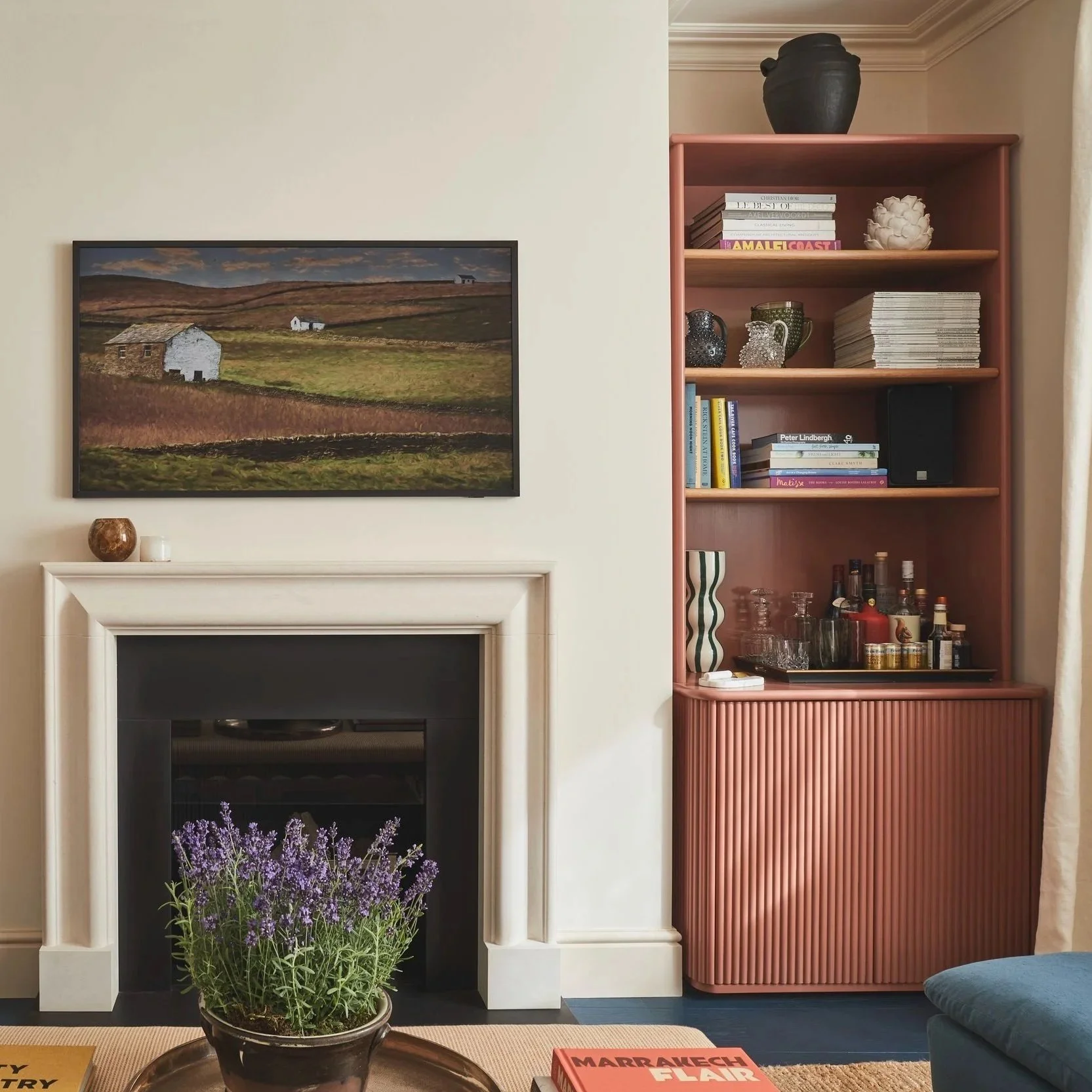 Sitting room with a fireplace, bespoke joinery, a painting of a countryside landscape, a pink bookshelf with decor and books, and a purple flower plant in a pot.