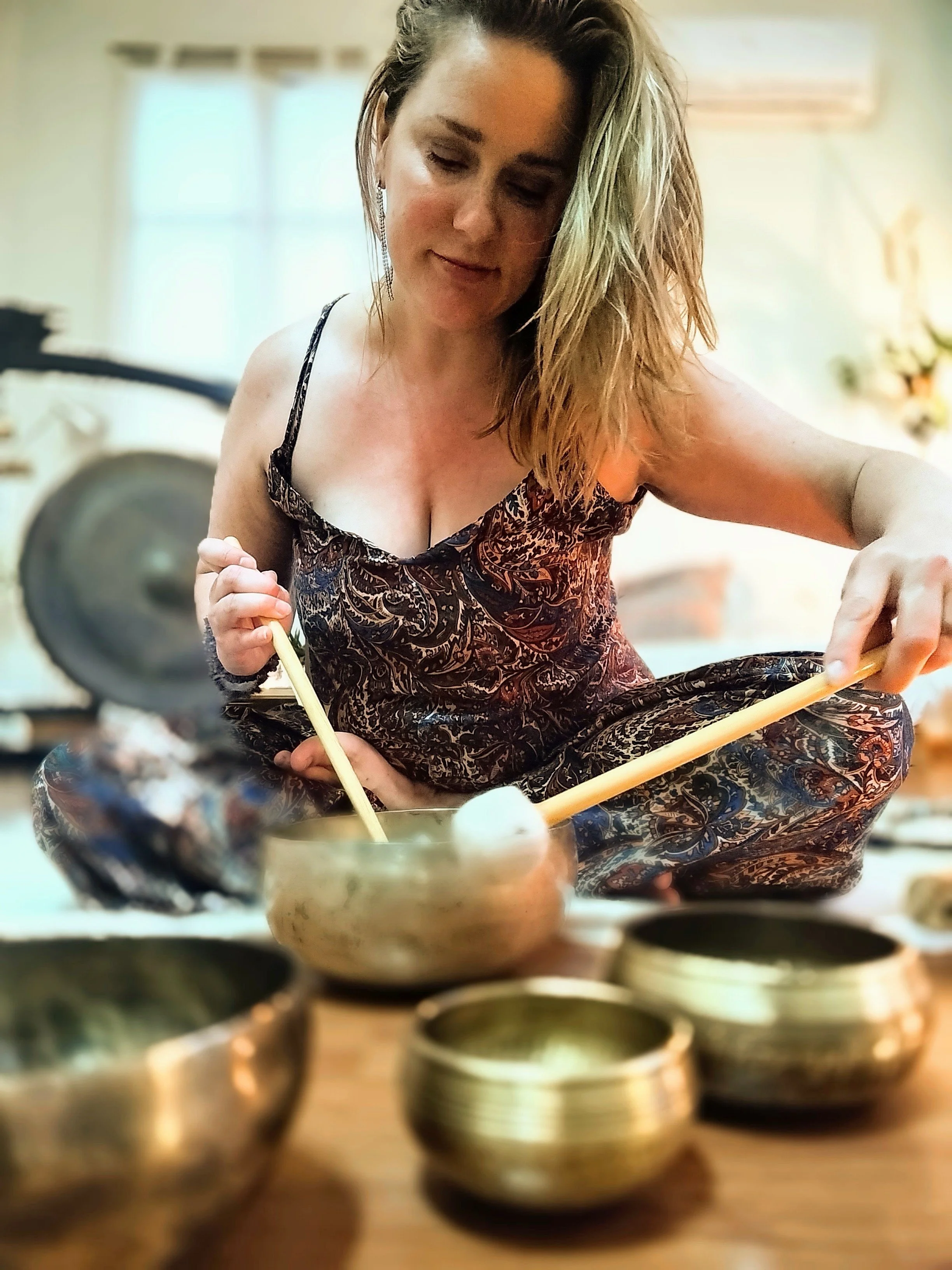 A woman with blonde, wavy hair, wearing a patterned dress, is sitting cross-legged in a room filled with sunlight, working with wooden sticks over a bowl. Sound bath Costa del Sol.