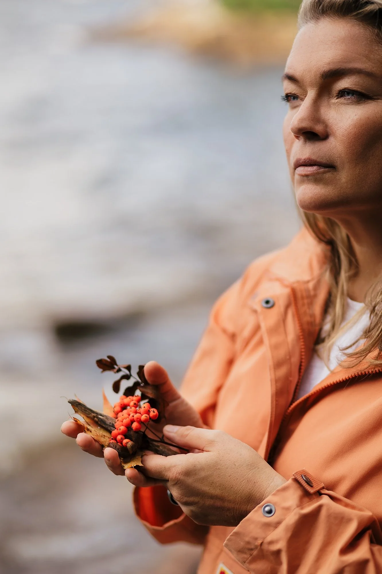 A woman wearing an orange jacket holding a small branch with orange berries and brown leaves.