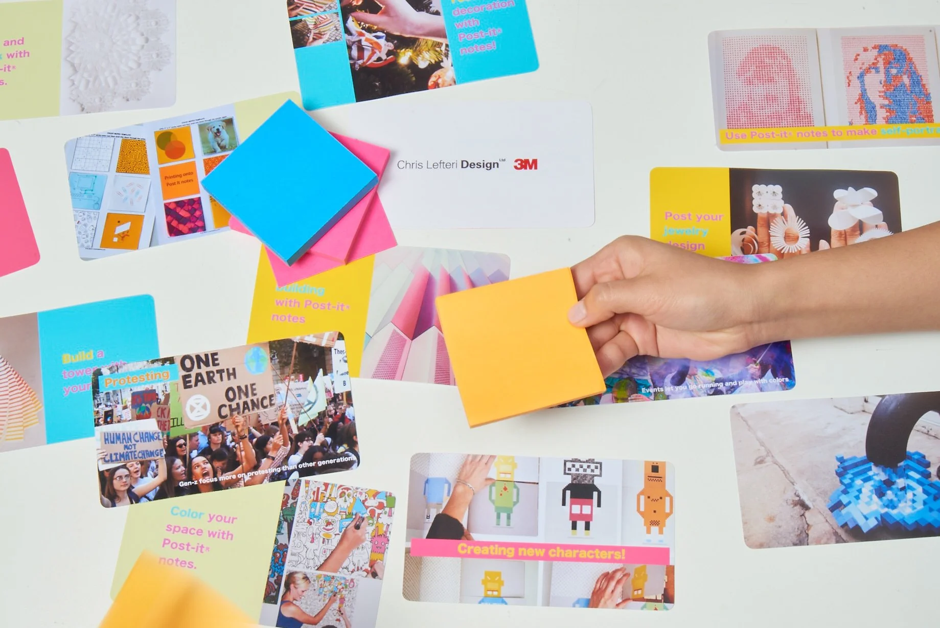Hand holding orange sticky note above a table with colorful Post-it notes and images. Various creative and artistic design projects using Post-it notes are displayed.