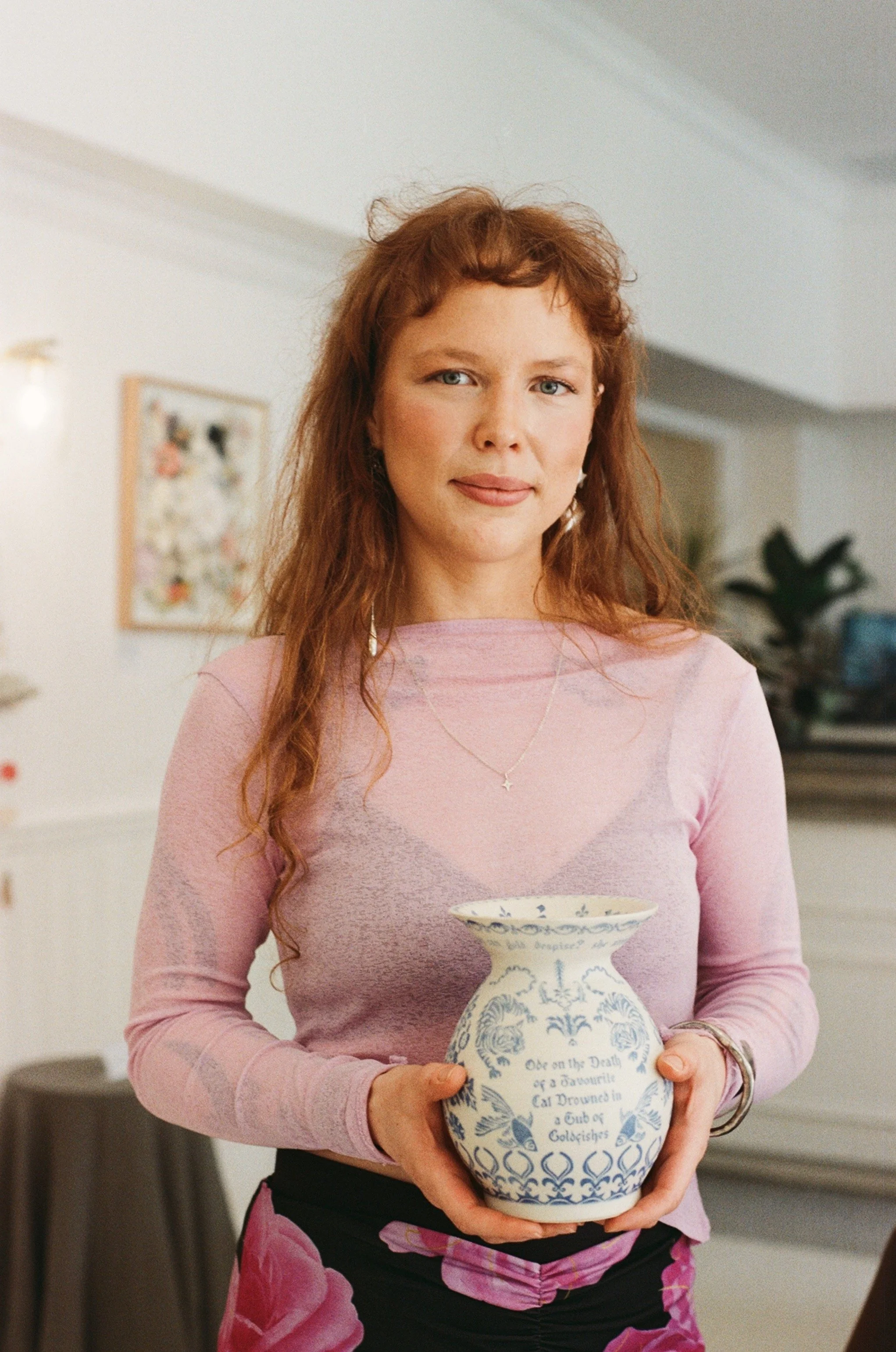 A woman with red hair holding a blue and white porcelain vase with text and floral patterns in a room with a framed picture on the wall and plants in the background.