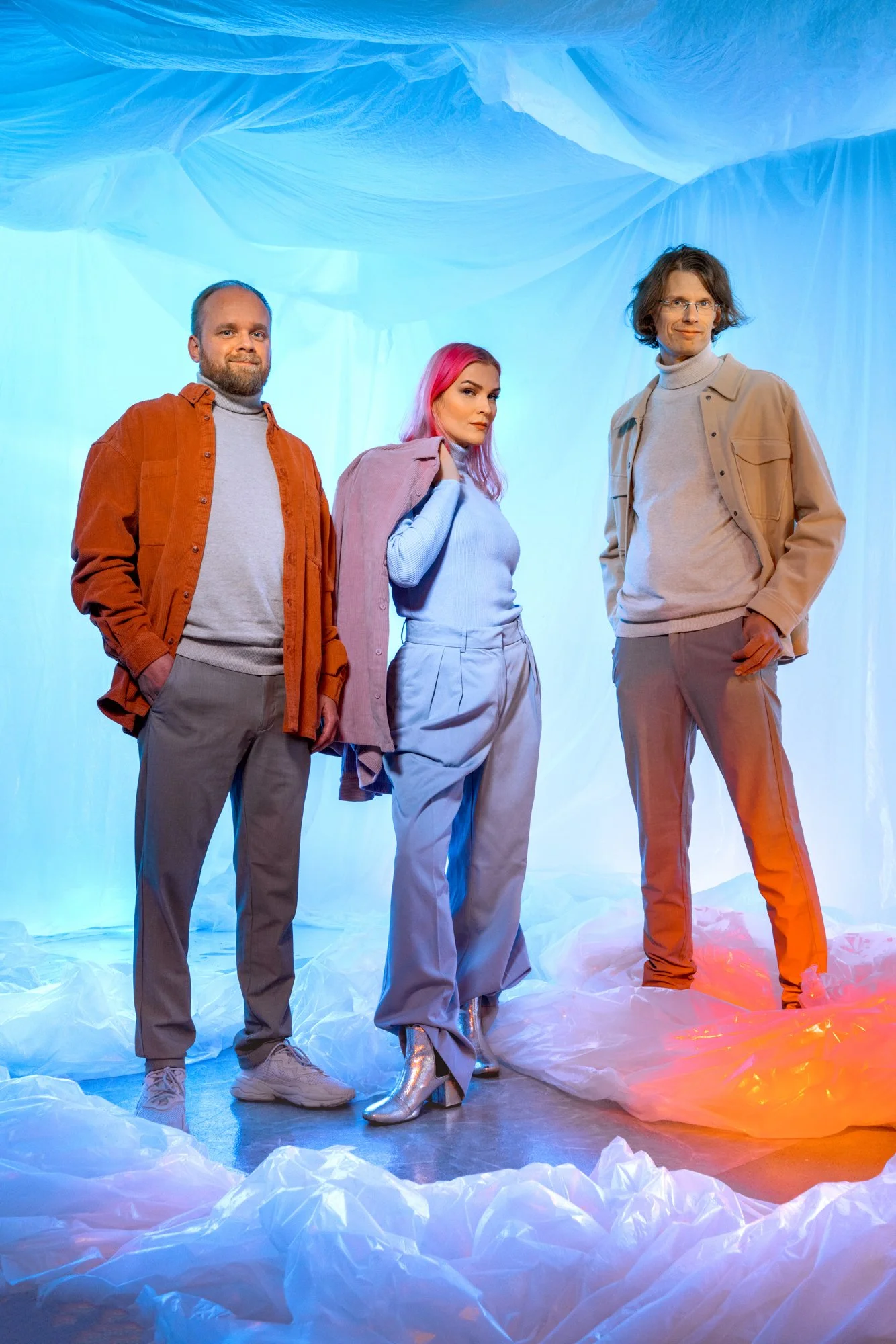 Three people standing in front of a blue backdrop with crumpled paper resembling clouds. They are dressed in casual clothing, with the woman in the middle wearing metallic silver boots, and the two men wearing neutral tones.