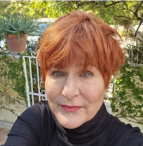 A woman with short red hair and blue eyes taking a selfie outdoors in front of greenery and a white gate.