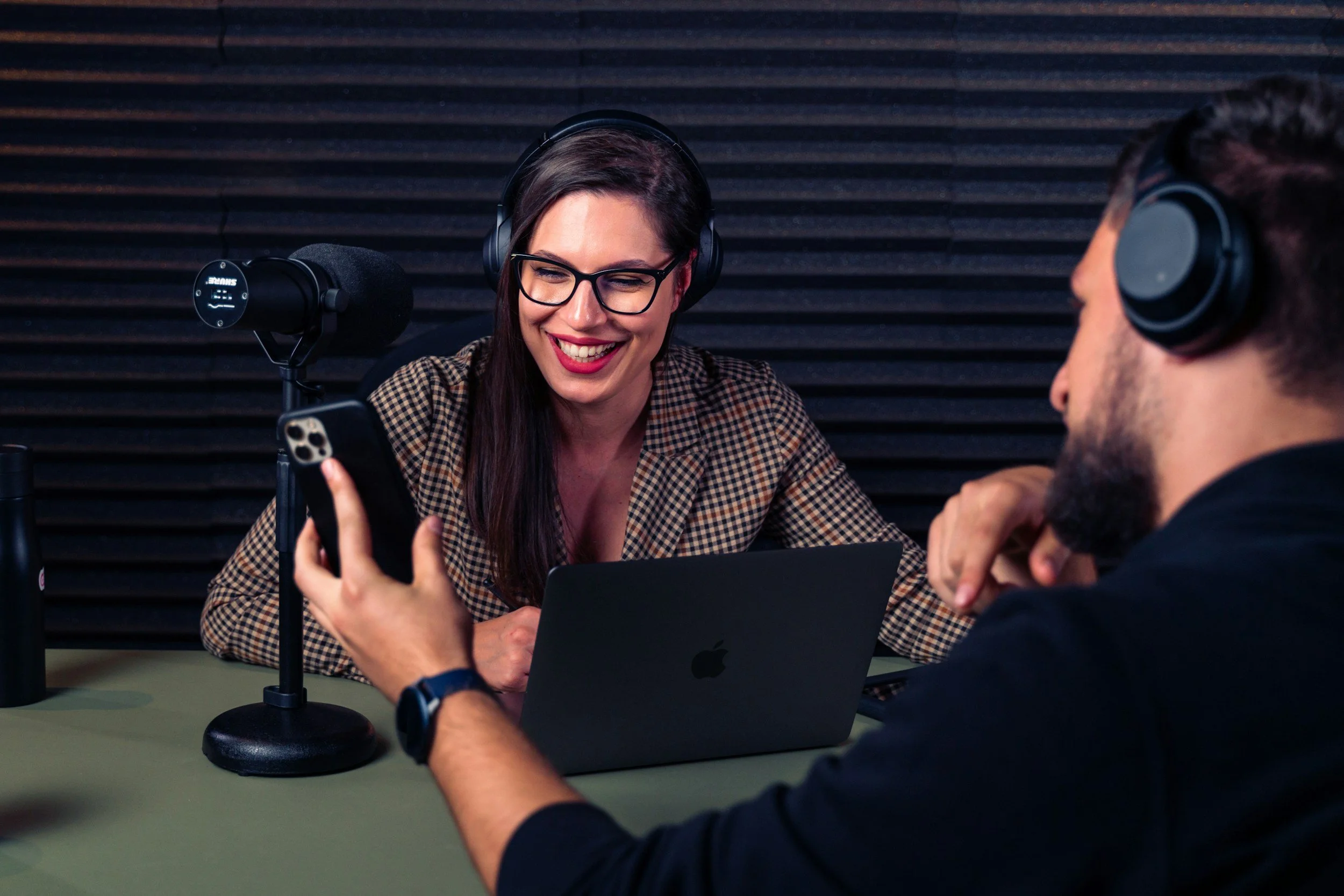 Two people wearing headphones, sitting at a radio station or recording studio, smiling and engaging with a smartphone, with a microphone and a laptop in front of them.