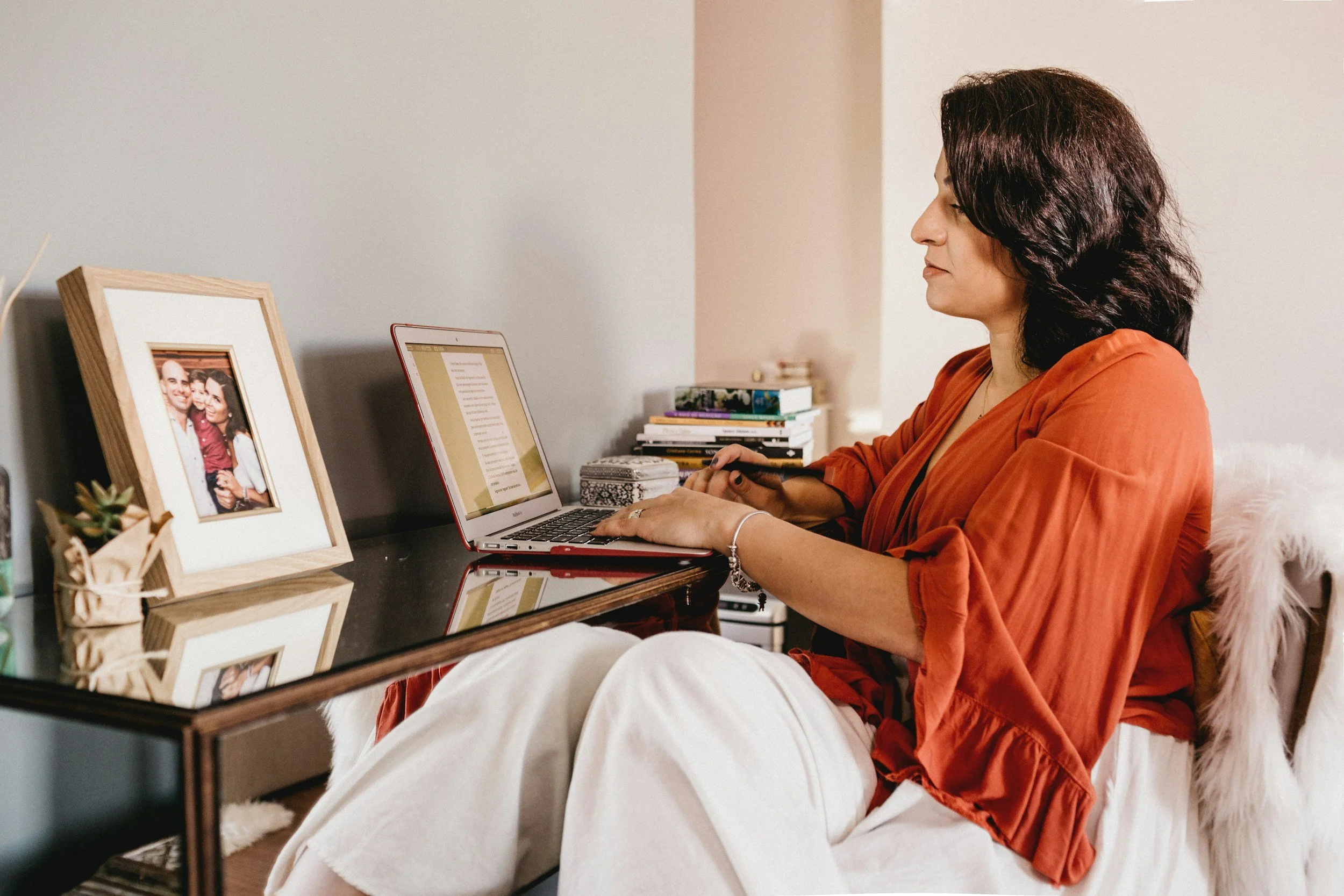 A woman with dark curly hair wearing an orange blouse, sitting at a desk with her hands on a laptop, in a cozy indoor setting with a stack of books and framed photographs on the desk.