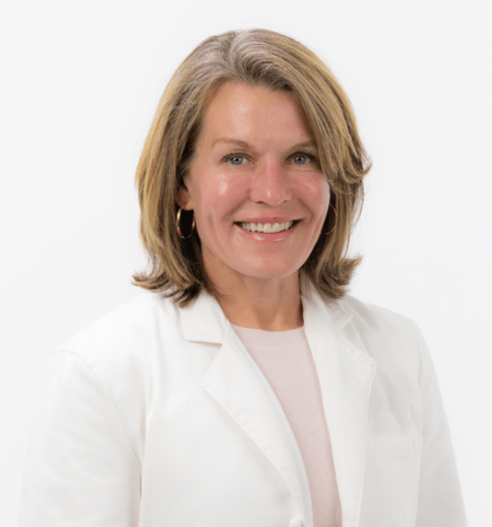 Portrait of a smiling middle-aged woman with shoulder-length blonde hair, wearing a white lab coat and hoop earrings, against a plain white background.