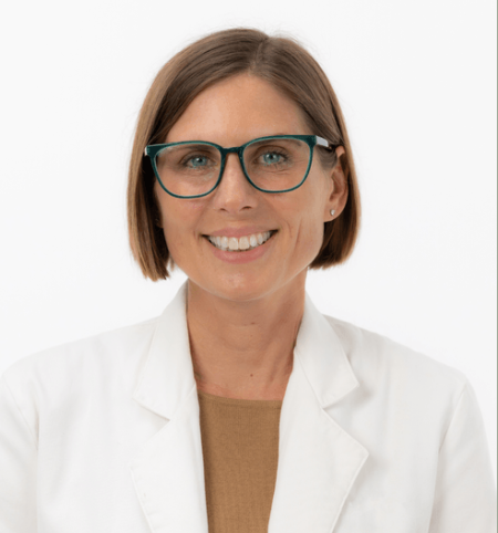 Smiling woman with short brown hair, wearing teal glasses and a white lab coat, against a plain white background.