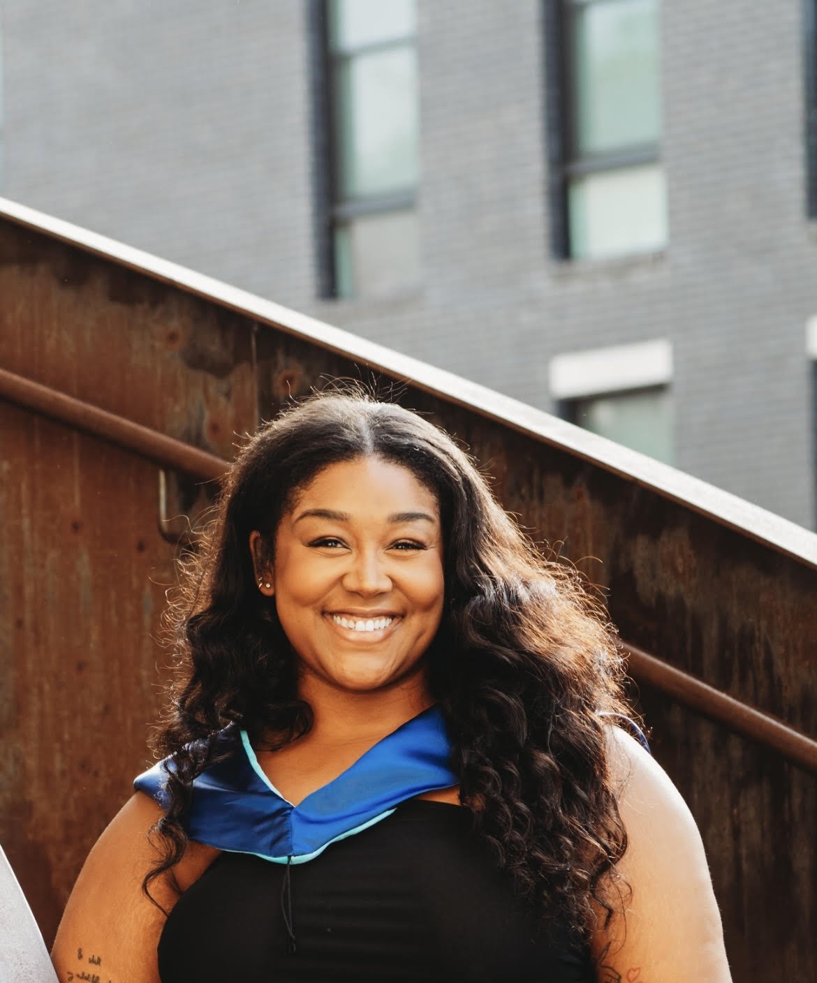 A young woman smiling outdoors with a wooden fence and a modern building in the background.