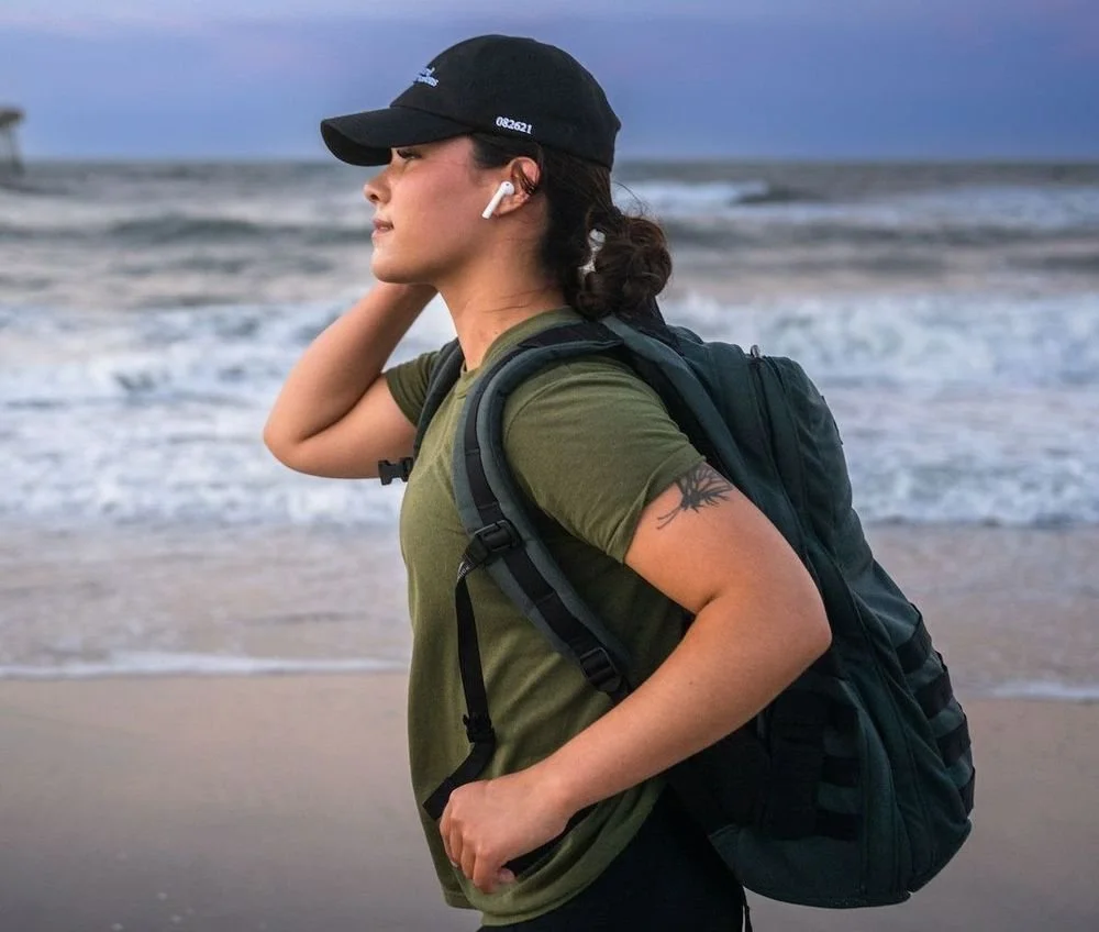 A woman with a tattoo on her arm wearing a black cap, green t-shirt, and wireless earbuds, carrying a backpack, standing on a beach with ocean waves in the background during dusk.