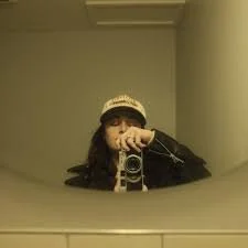 Person taking a mirror selfie with a camera, wearing a cap, in a room with beige walls.