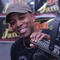 Smiling young man wearing a gray cap and black shirt, holding a microphone, in front of a colorful background.