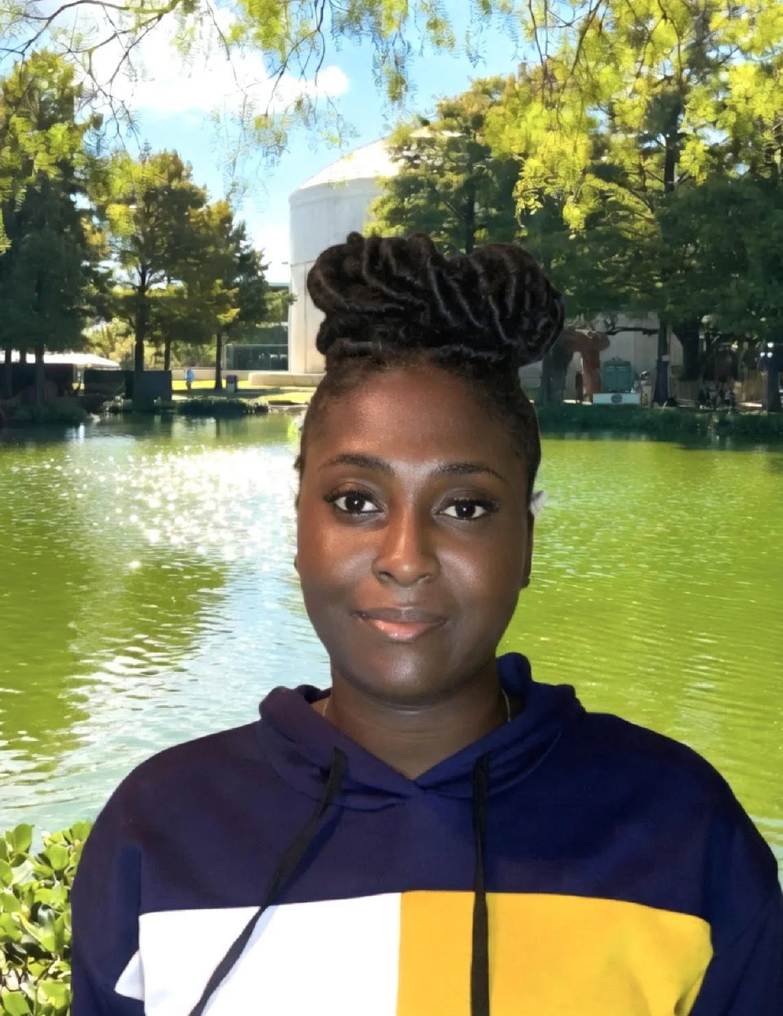 A woman with dark skin and hair styled in large twists in a bun, smiling and wearing a navy blue hoodie with a white, yellow, and black design. She is outdoors near a body of water, with trees and a white building in the background on a sunny day.