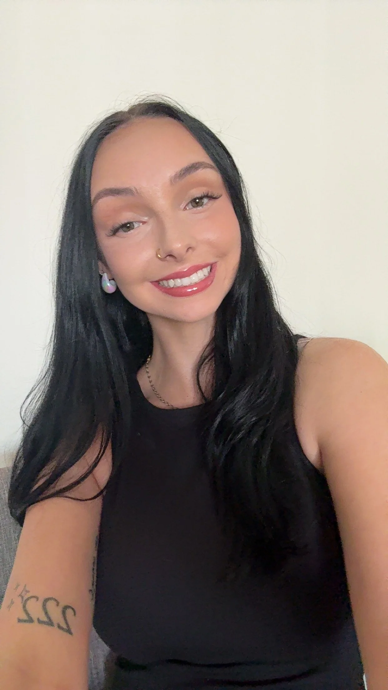 A young woman with long black hair takes a selfie against a plain white wall. She has a nose ring, earrings, and wears a black sleeveless top. She is smiling with red lipstick and shows a tattoo on her left arm.