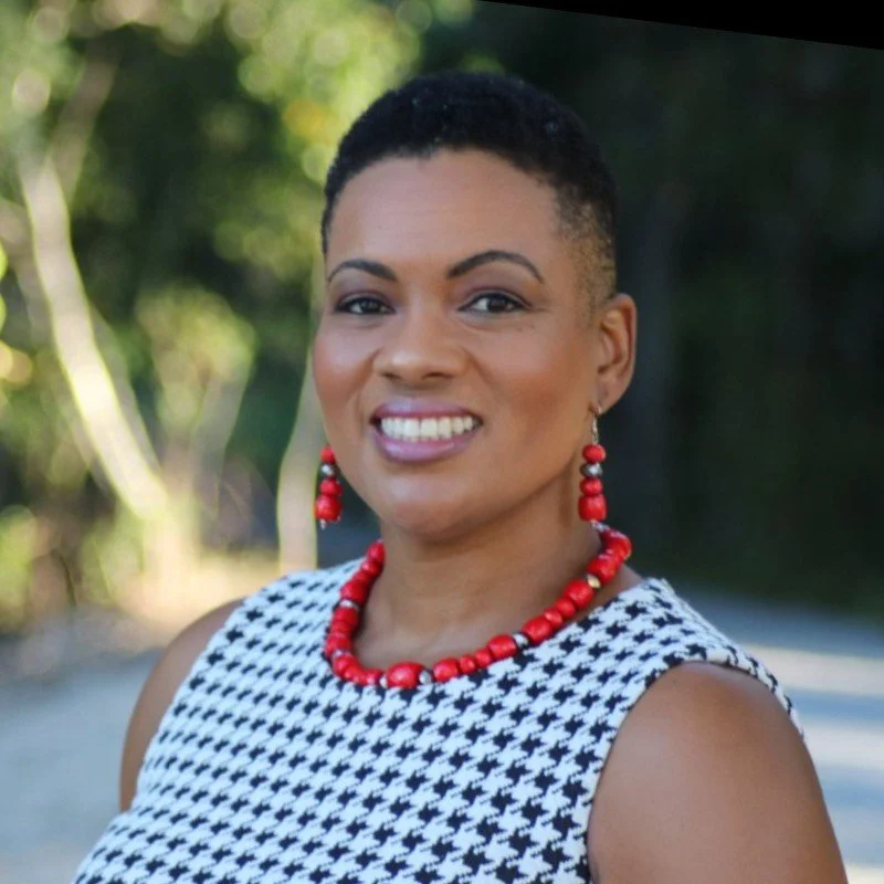 A woman with short dark hair smiling outdoors, wearing a black and white houndstooth sleeveless top and red jewelry against a blurred nature background.