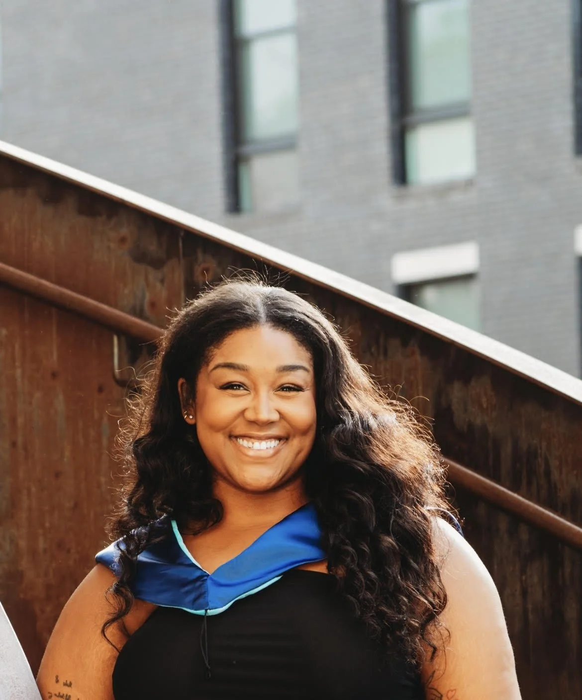 A young woman with dark, curly hair smiling outdoors, wearing a black sleeveless top and a blue scarf around her neck. There is a wooden railing and a brick building with large windows in the background.