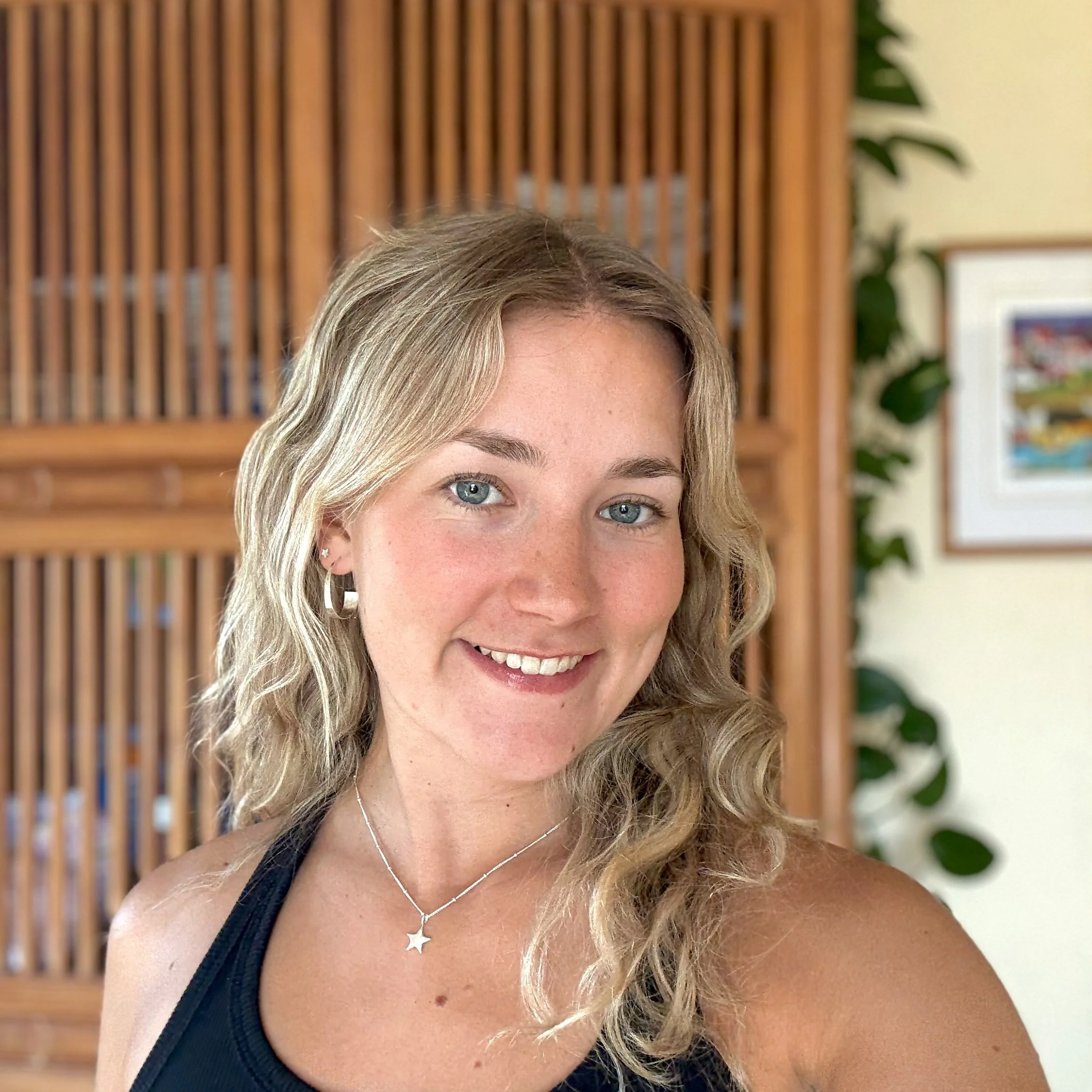 Headshot of young blonde silversmith Georgia wearing her spiral hoop earrings and a silver star necklace. In the background is a bamboo screen and plants.