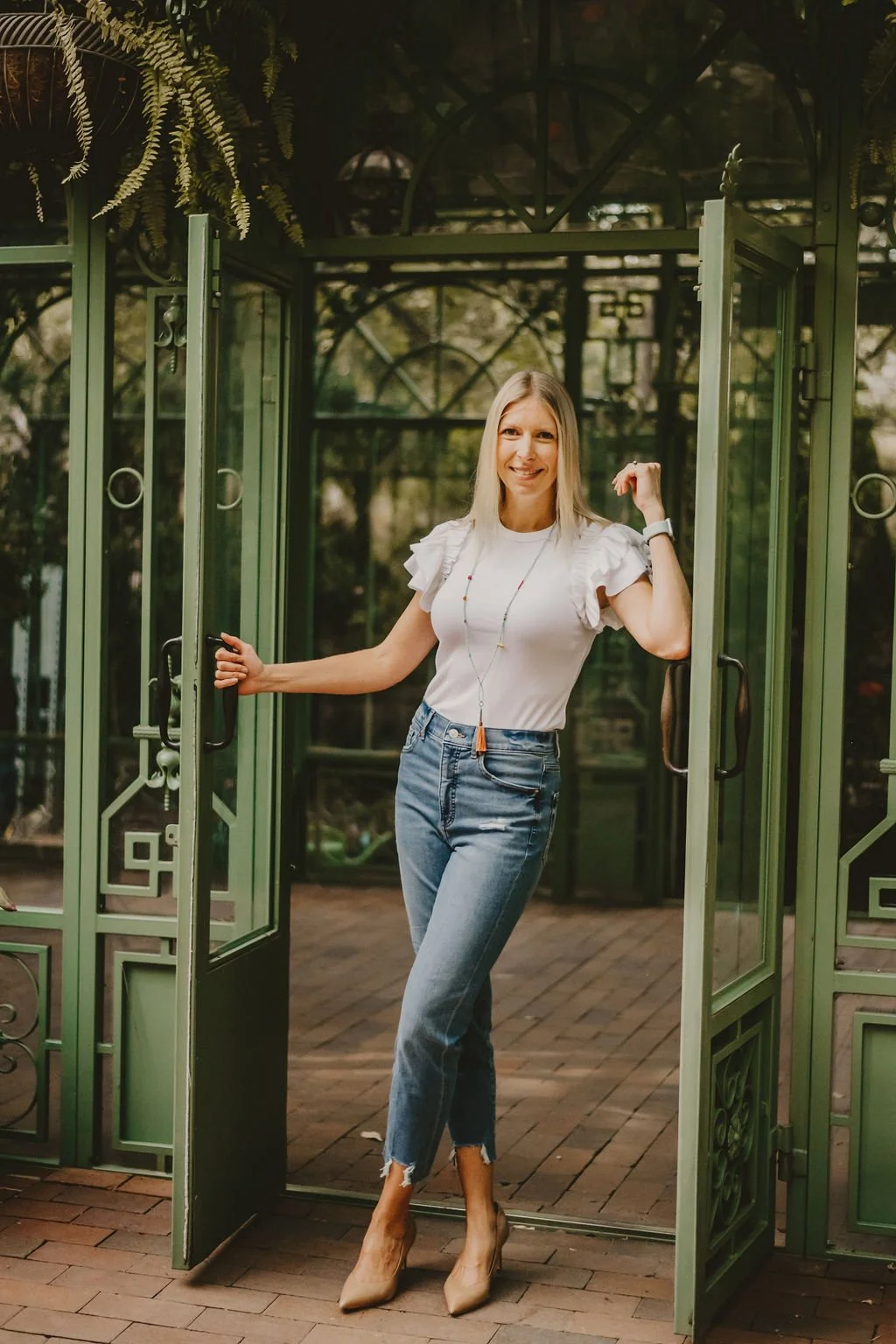 A woman standing in the doorway of a green metal greenhouse, smiling and posing with one hand on the door handle and the other flexed.