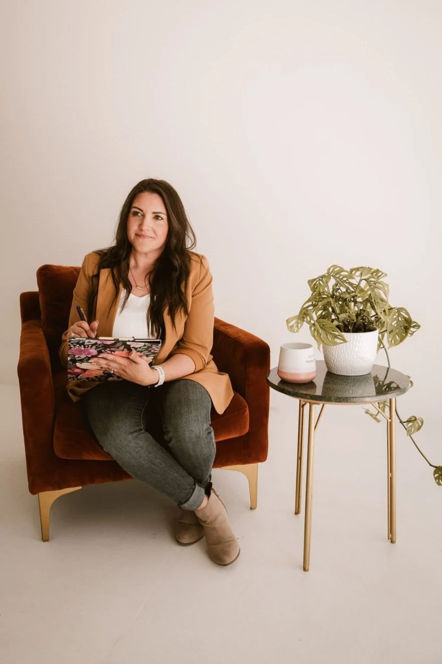A woman with dark hair sitting in a burnt orange velvet armchair, holding a floral planner, on a white background. There is a small black table with a potted plant and a modern white and pink candle holder beside her.