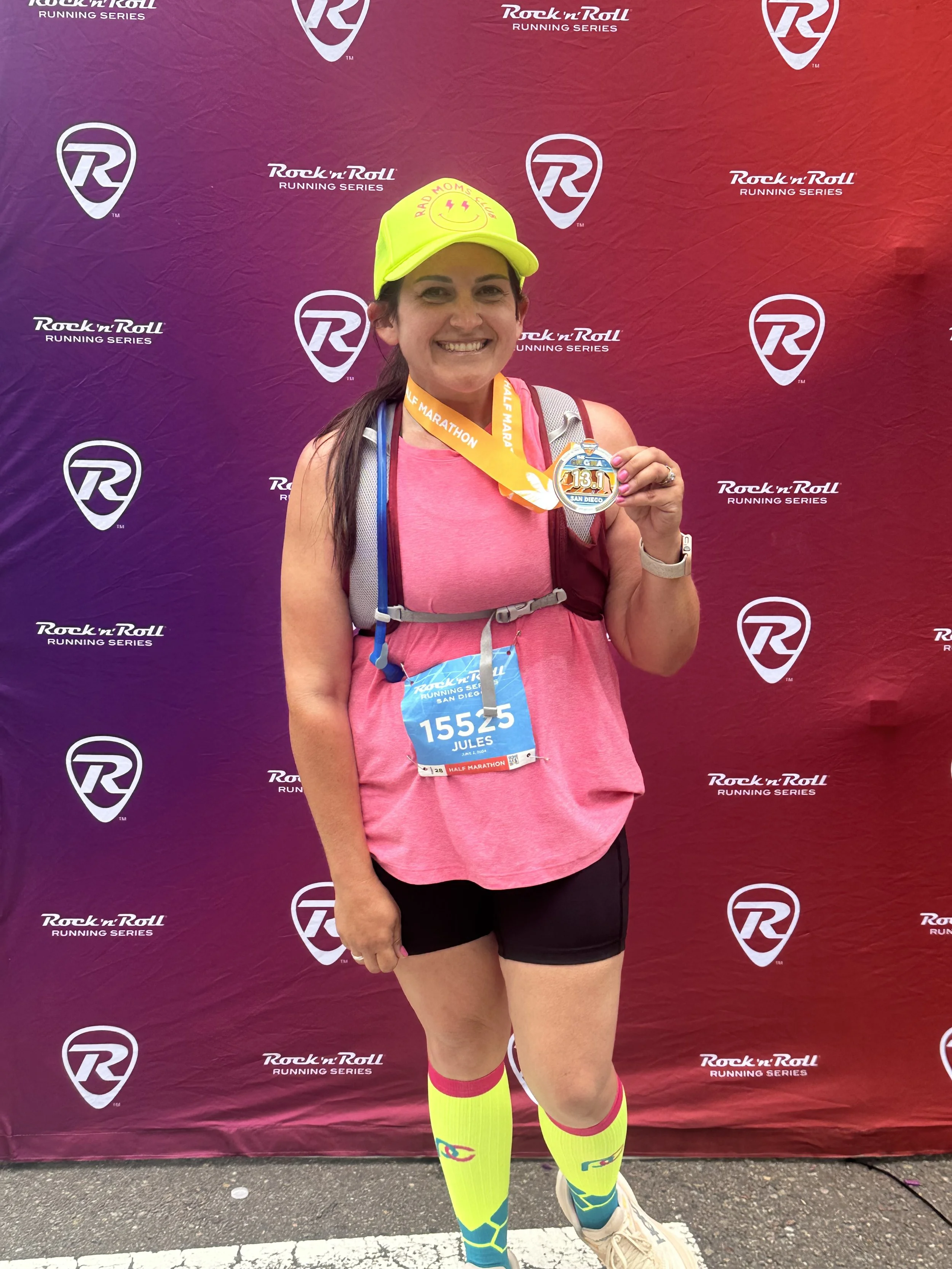 A woman standing in front of a purple background with the Rock 'n' Roll Running Series logo, wearing a pink athletic top, black shorts, neon yellow and blue running socks, a neon yellow cap, and holding a medal. She is smiling and has a race bib with the number 15525 and the name Jules.