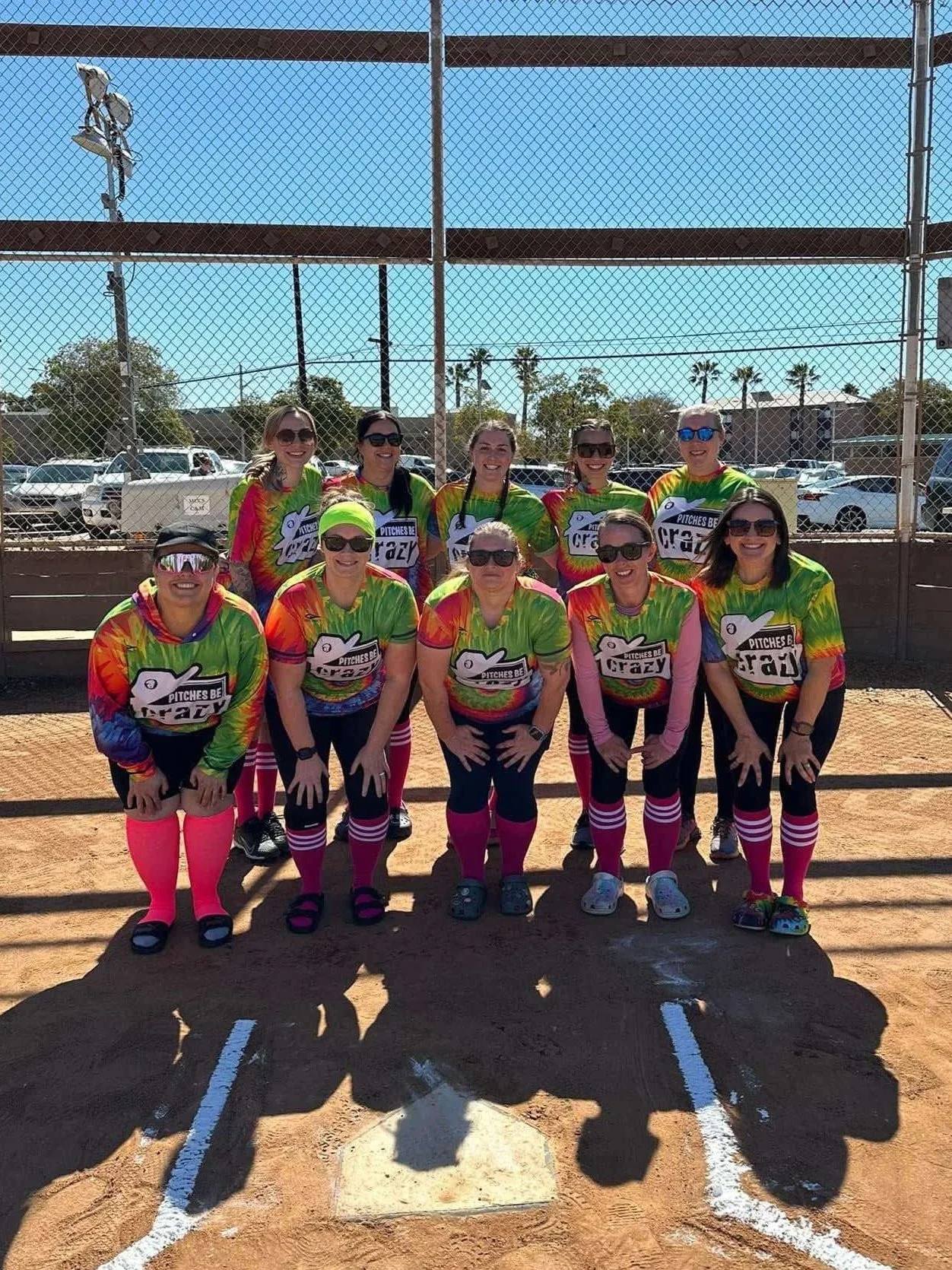 Group of nine women in rainbow-colored softball uniforms posing on a dirt field with chain-link fence behind them and parked cars in the background.