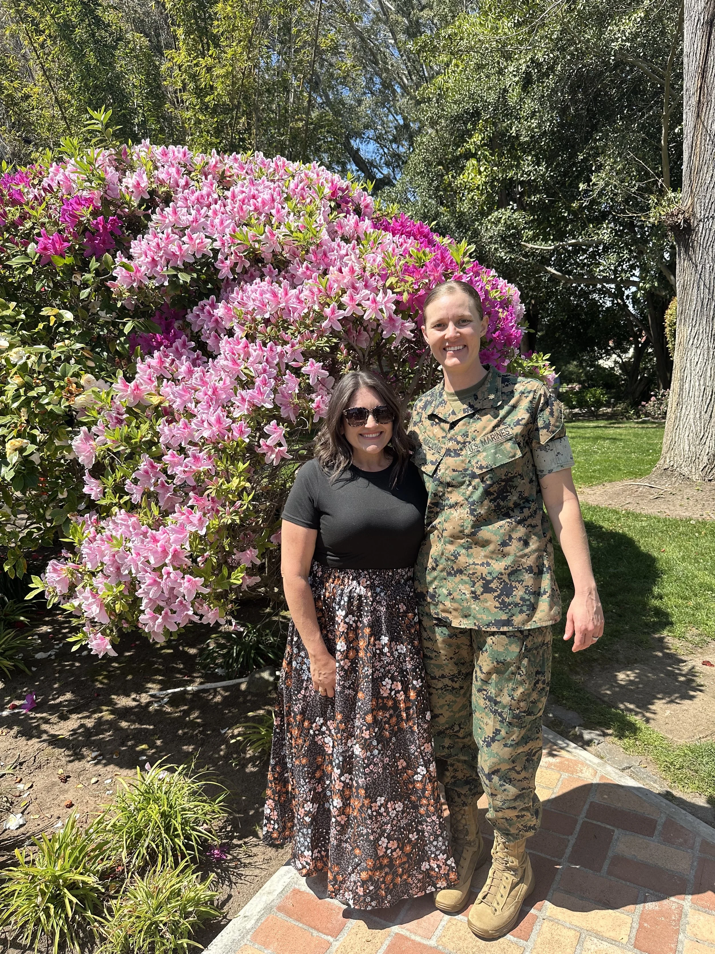 A woman in a black top and floral skirt standing next to a woman in military uniform, both smiling, in front of a large bush of pink and purple flowers, outdoors on sunny day.