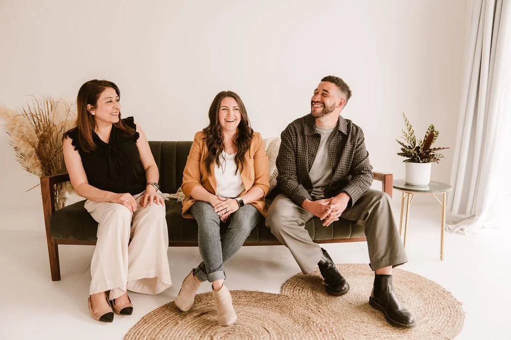 Three people sitting on a black sofa, smiling and laughing, with a potted plant and dried pampas grass in the background, in a bright, minimalist living room.