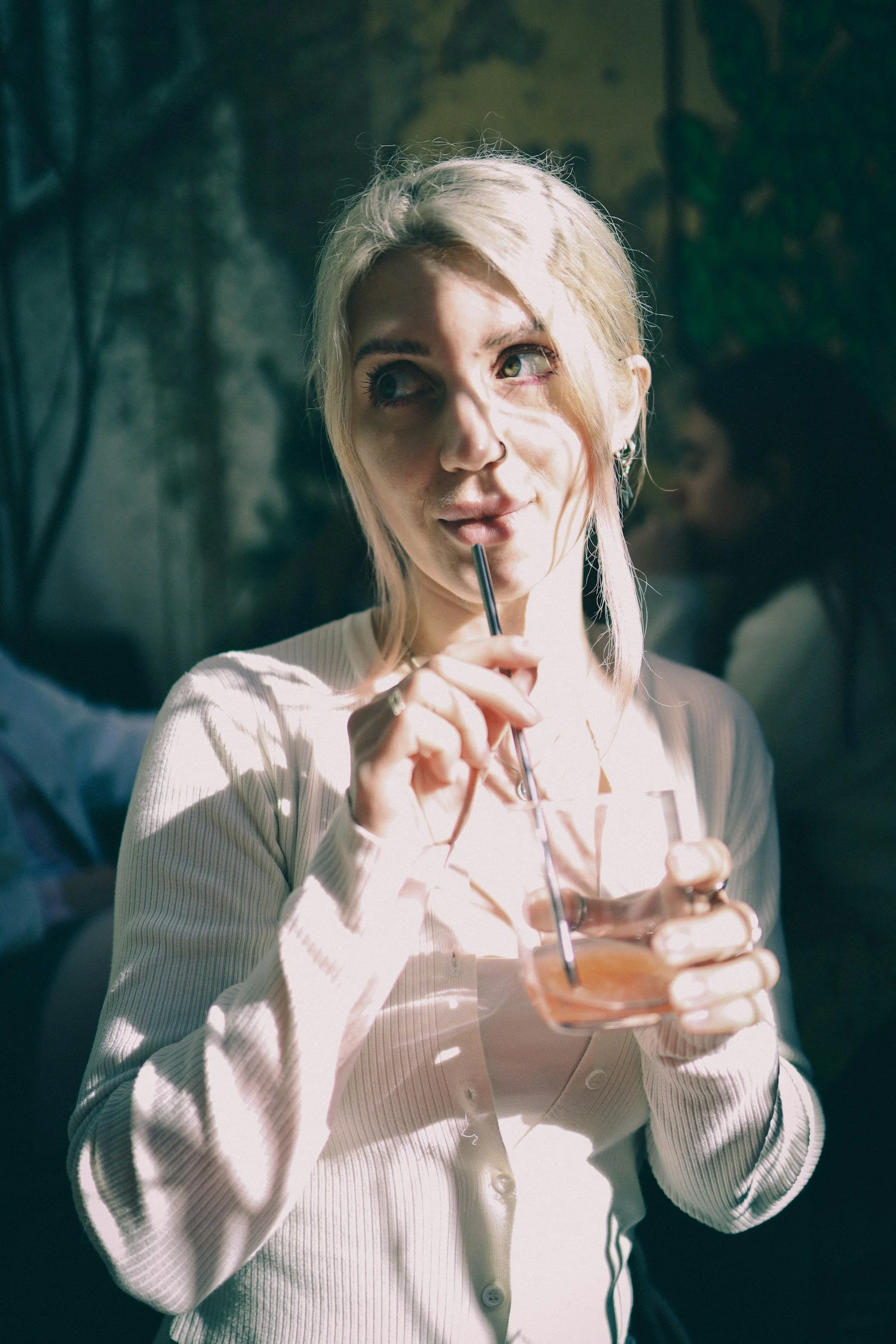 Blonde woman holding a glass of water with a straw in it, held up to her mouth, in sunlit surroundings