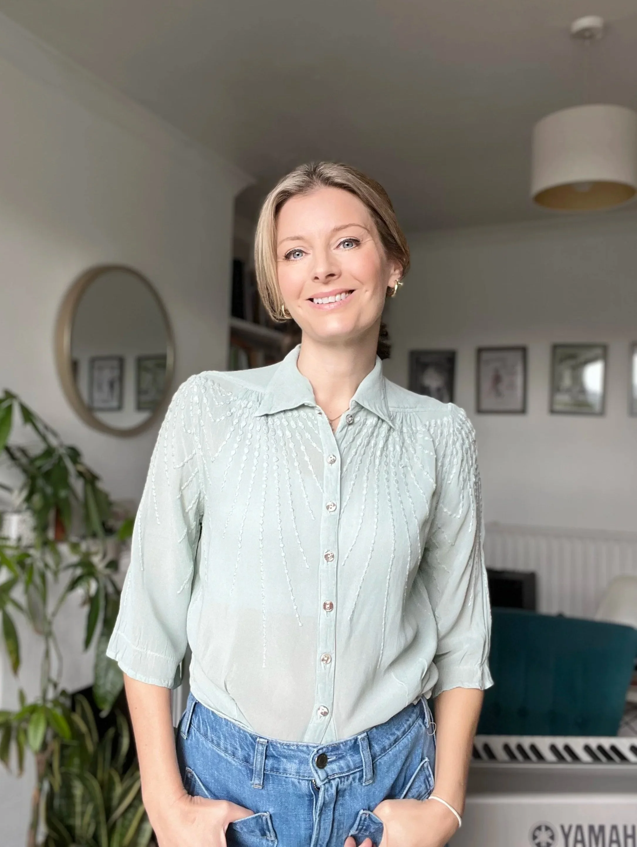 Laura Boon singing teacher, standing in the music teaching room in front of her keyboard, with her hands in her pockets and smiling at the camera. Her hair is in a ponytail and she is wearing jeans and a pale turquoise blouse.