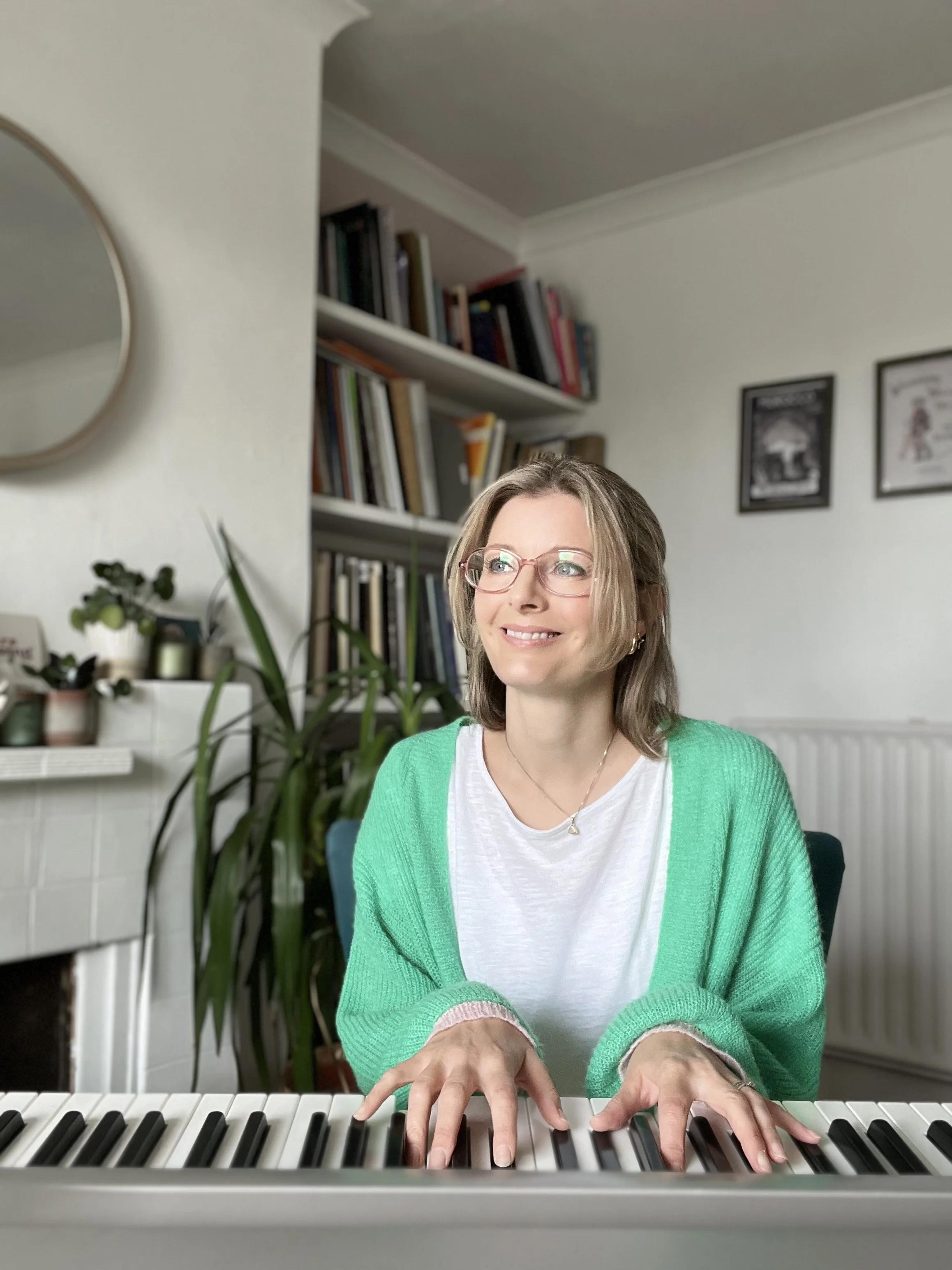 Smiling Laura singing teacher wearing glasses and a green cardigan playing an electronic keyboard in her home music studio