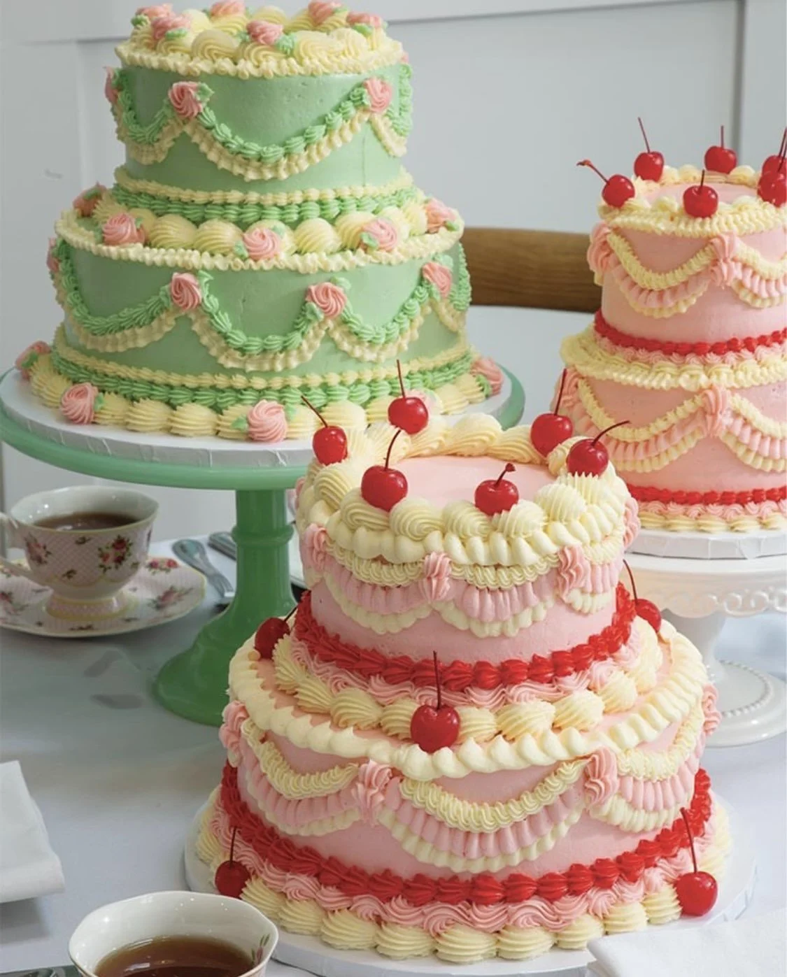Three decorated layered cakes with pink, white, and green frosting and cherry toppings, displayed on cake stands. There is also a tea cup with tea on the table.