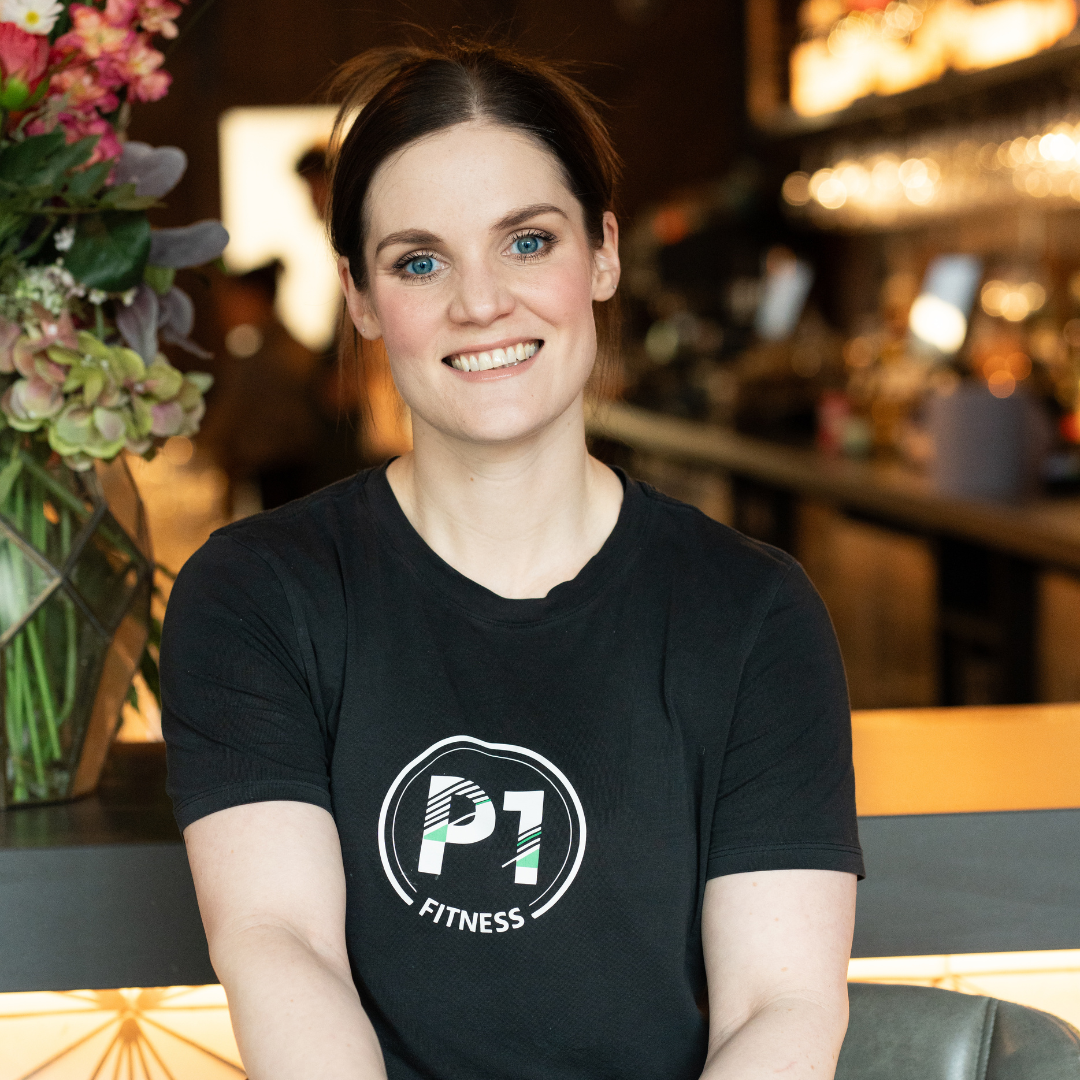 Young woman with short dark hair and blue eyes smiling, sitting indoors at a place that appears to be a gym or fitness center, wearing a black t-shirt with the logo 'P1 Fitness'.