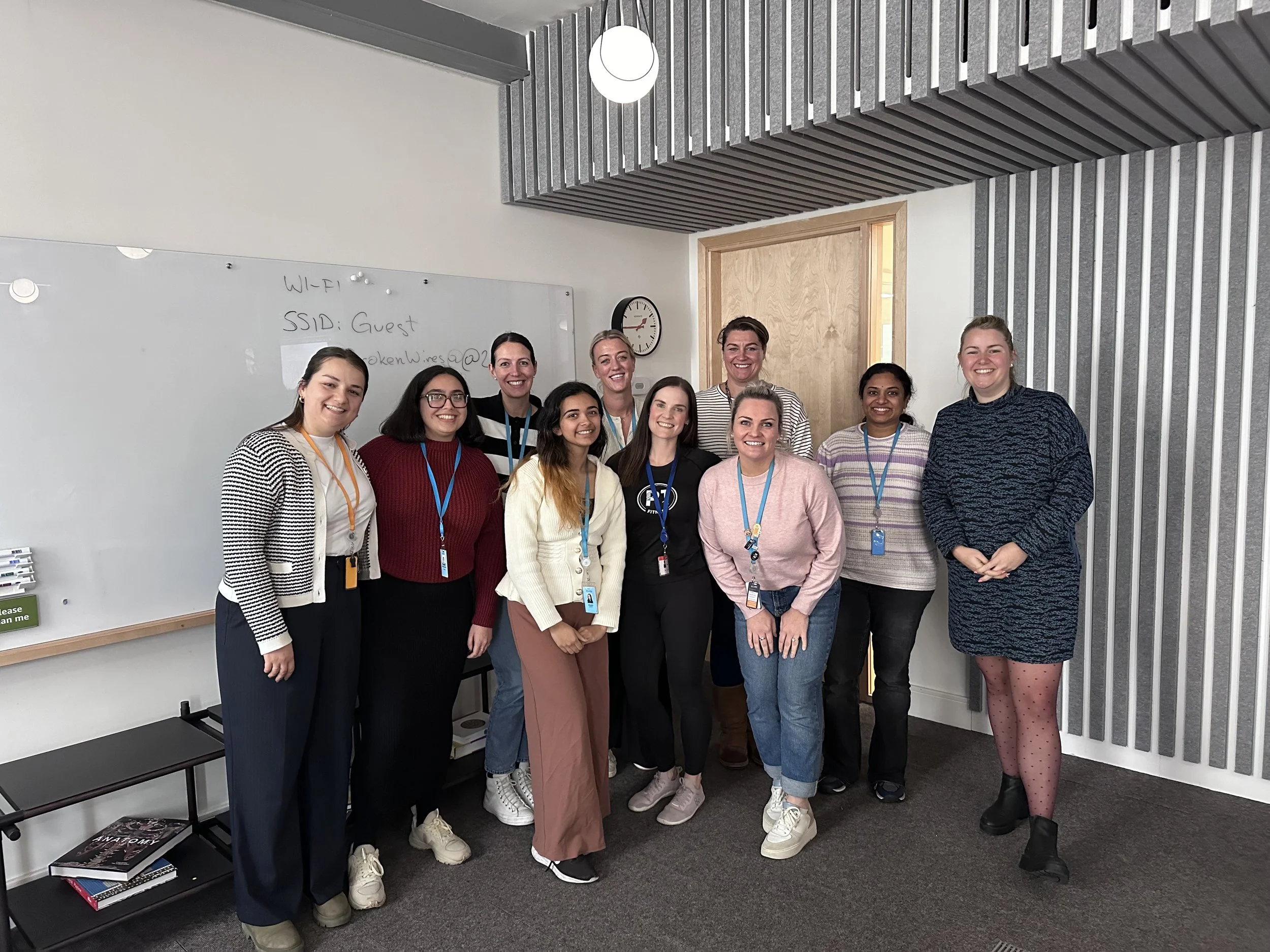Group of ten women standing together indoors, smiling for the camera. They are in a room with a whiteboard on the wall and a door behind them.