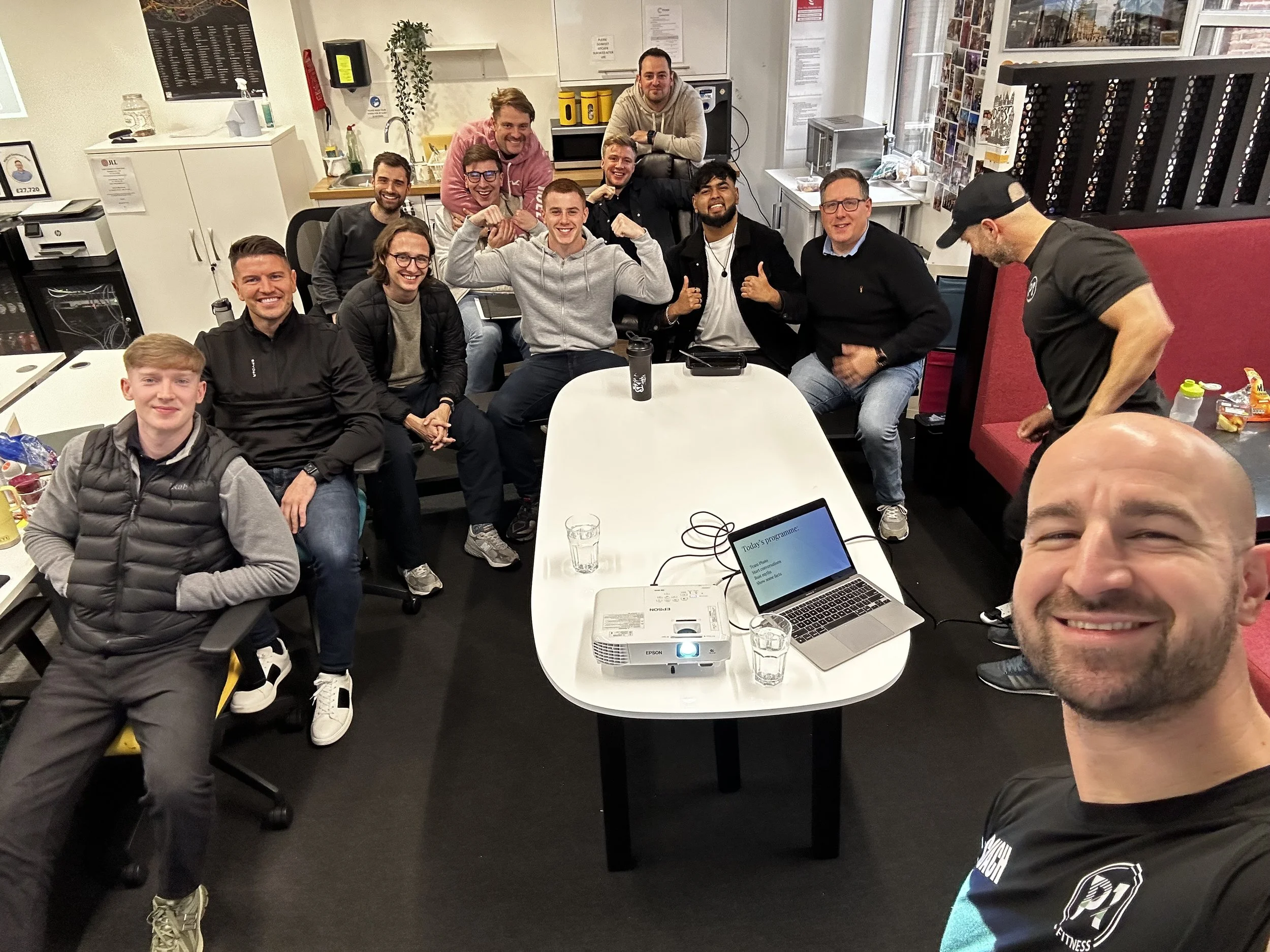 Group of people gathered in a casual office setting, taking a selfie. The group is smiling and posing around a table with a laptop and a projector, with some individuals standing and others sitting, in a well-lit room with office supplies and a window in the background.