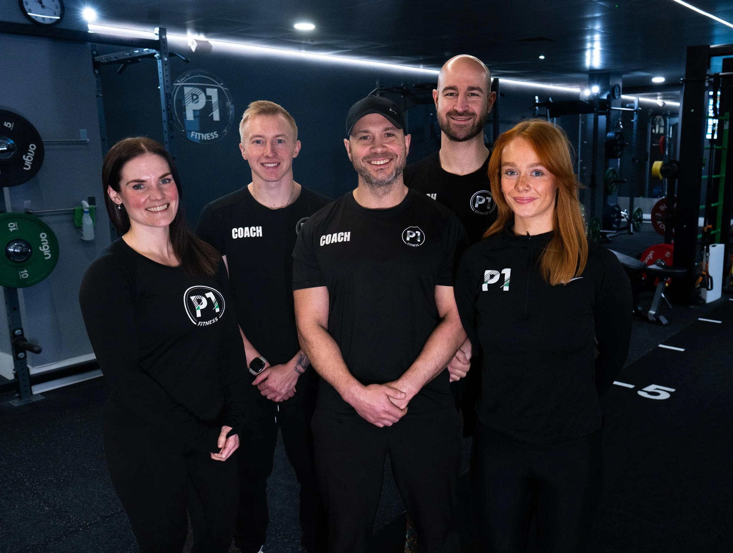Group of five fitness trainers in black athletic clothing standing in a gym with workout equipment in the background.