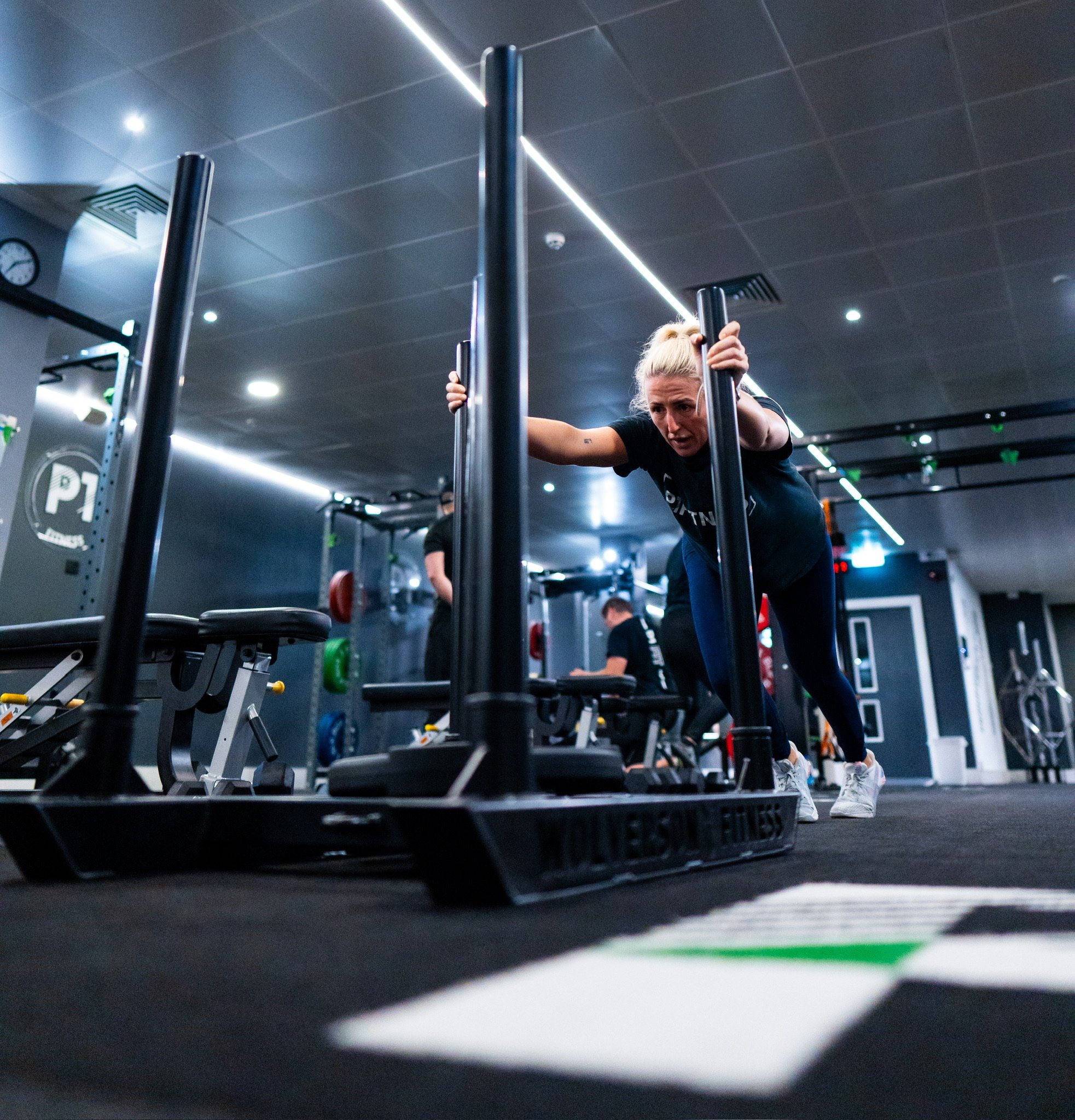 A woman in athletic clothing is pushing a sled weighted with weights in a gym, with other people working out in the background.