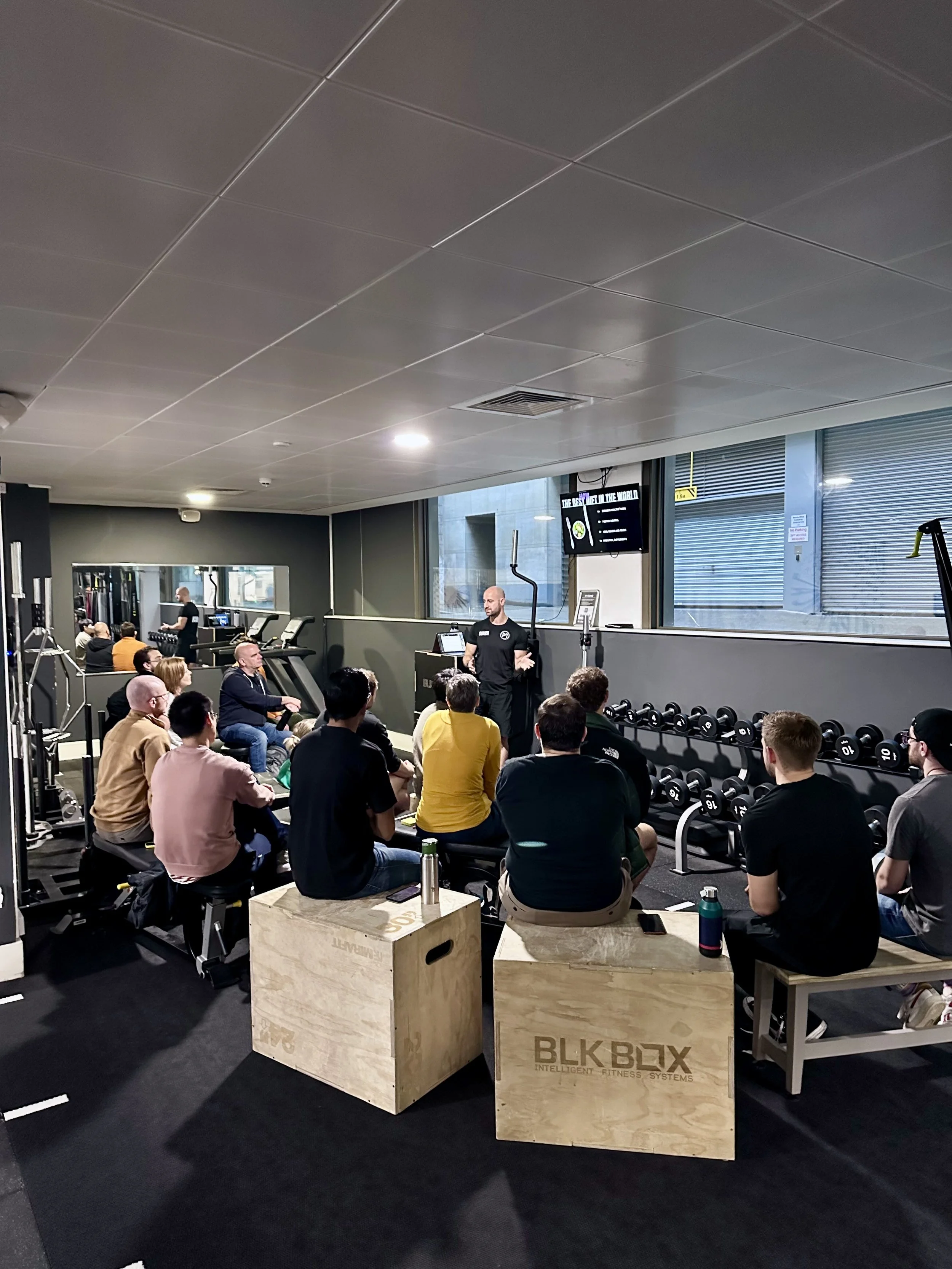 A fitness class or seminar taking place in a gym with an instructor leading a session while attendees sit on benches and boxes, facing a screen displaying information.