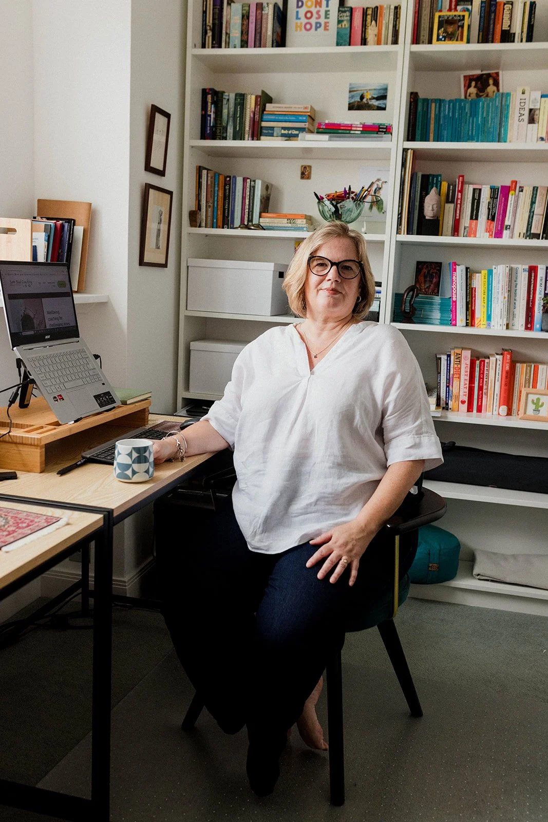 Sam with glasses and a white blouse sitting in a home office, with a bookshelf filled with books behind her, a desk with a laptop, coffee mug, and various office supplies.