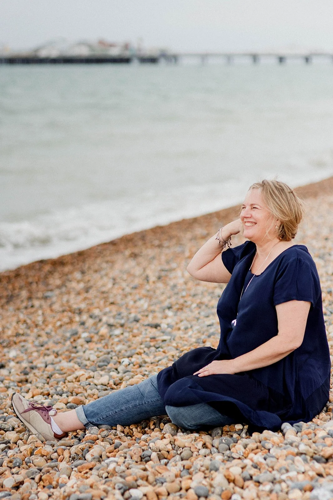 Sam is sitting on a pebble beach smiling and looking to the right, with the ocean and Brighton pier in the background.