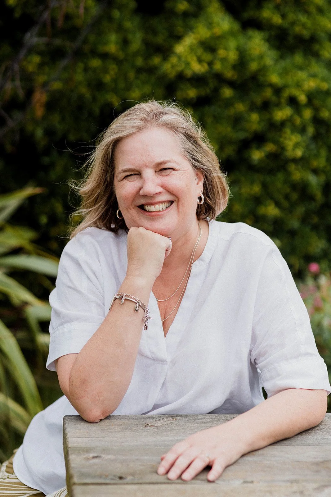 Sam with blonde hair and hoop earrings is smiling and sitting at a wooden table outdoors, with greenery in the background.