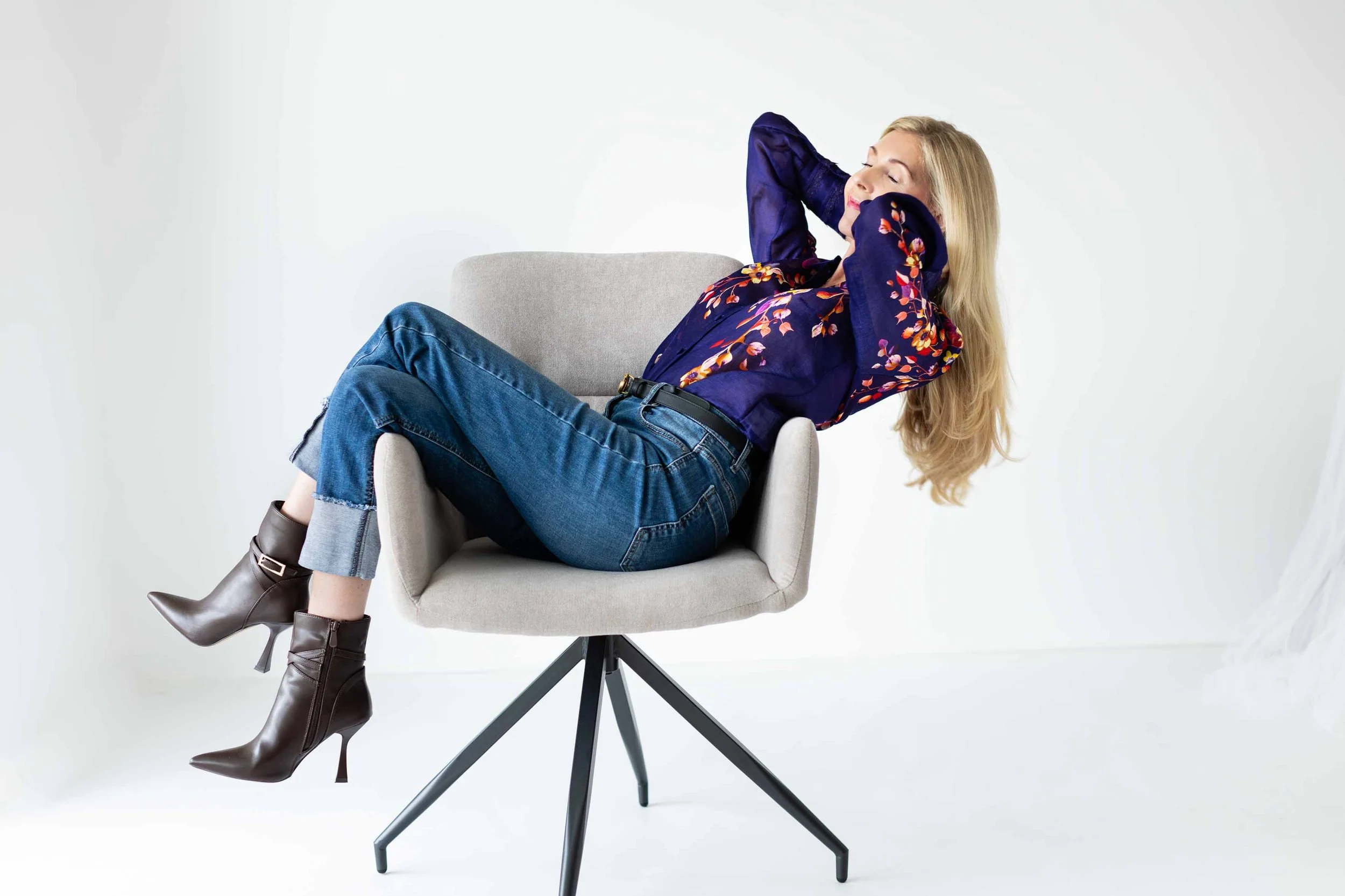 Frau sitzt entspannt auf einem modernen, grauen Sessel, trägt eine blaue Bluse mit Blumenmuster, Jeans und braune High Heels, hat die Hände im Haar und schließt die Augen.