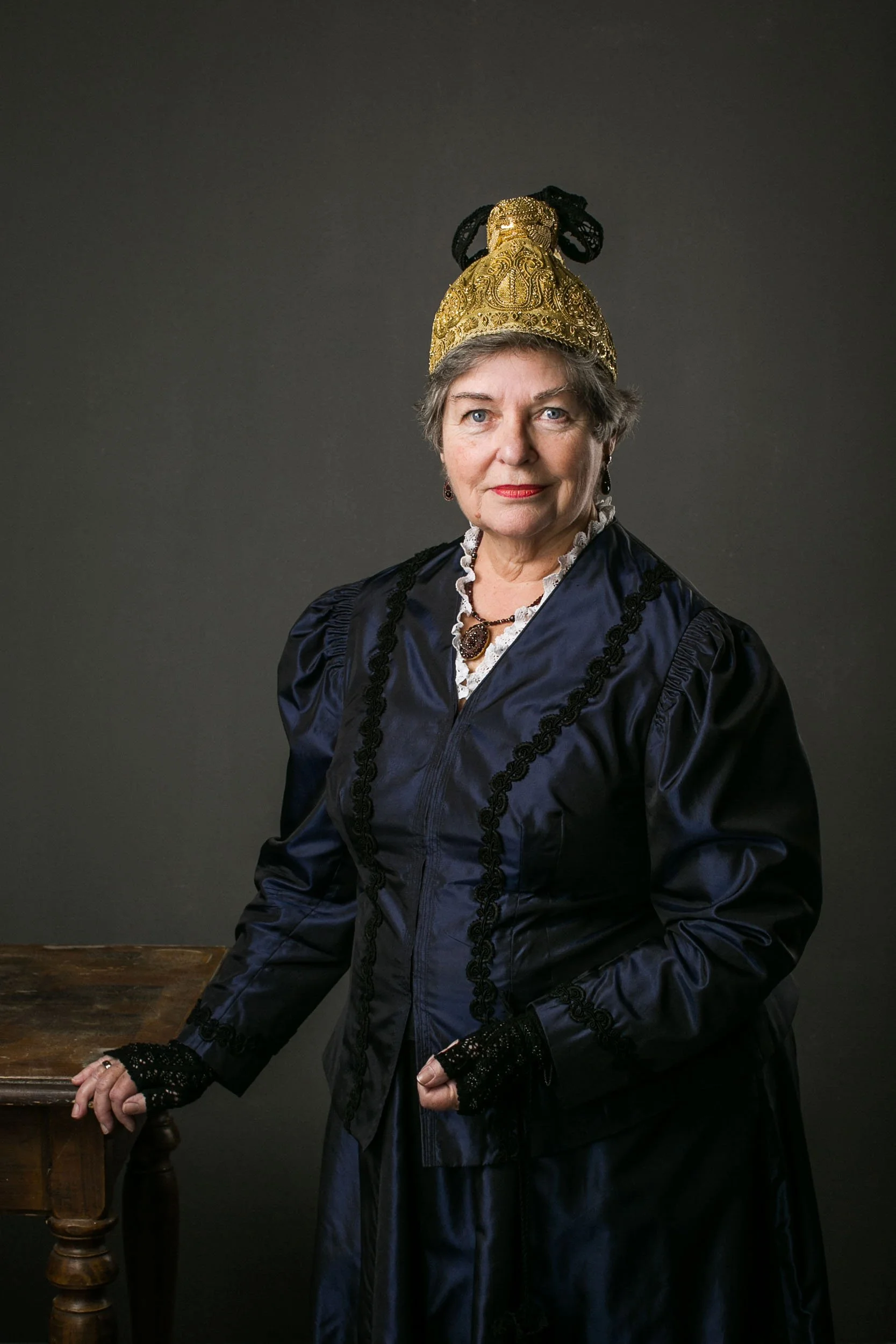 Porträt einer älteren Frau in viktorianischer Kleidung mit einer goldenen, verzierten Kappe und schwarzen Handstulpen vor dunklem Hintergrund.