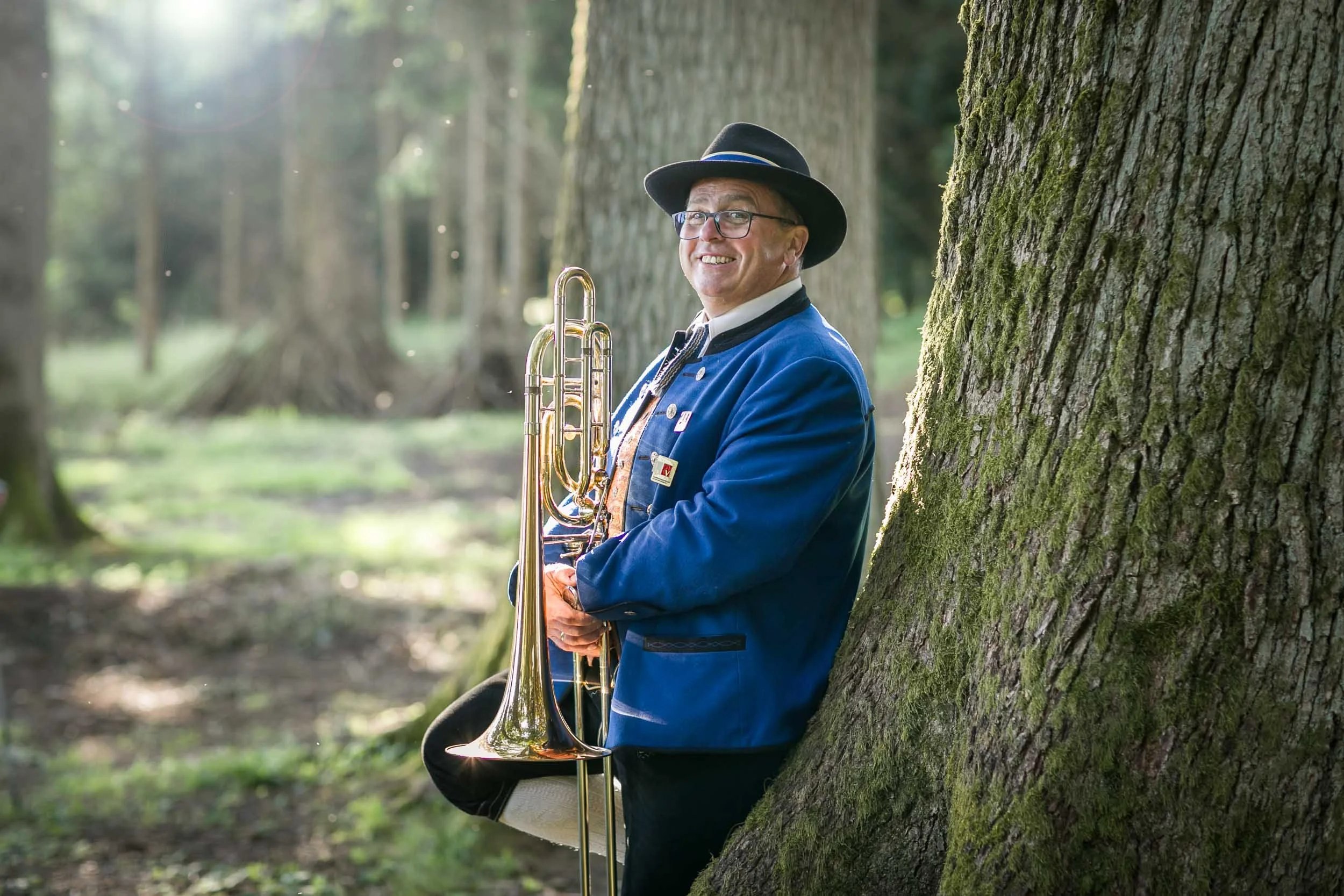 Ein Mann in traditioneller Trachtenkleidung mit Hut, Brille und einer Trompete, der im Wald neben einem Baum steht und lächelnd in die Kamera schaut.