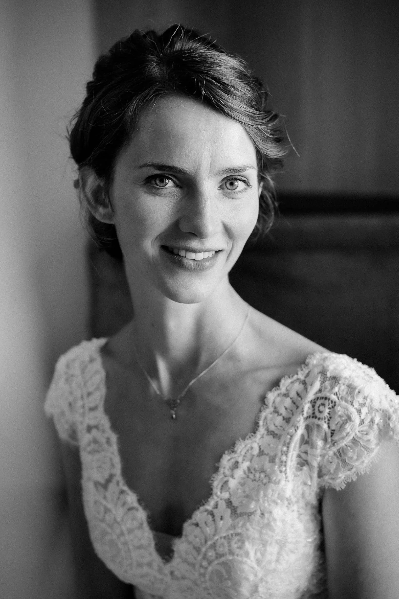 Schwarze und weiße Nahaufnahme einer lächelnden Frau in einem Brautkleid mit Spitze, die Schatten an der Wand lehnt.