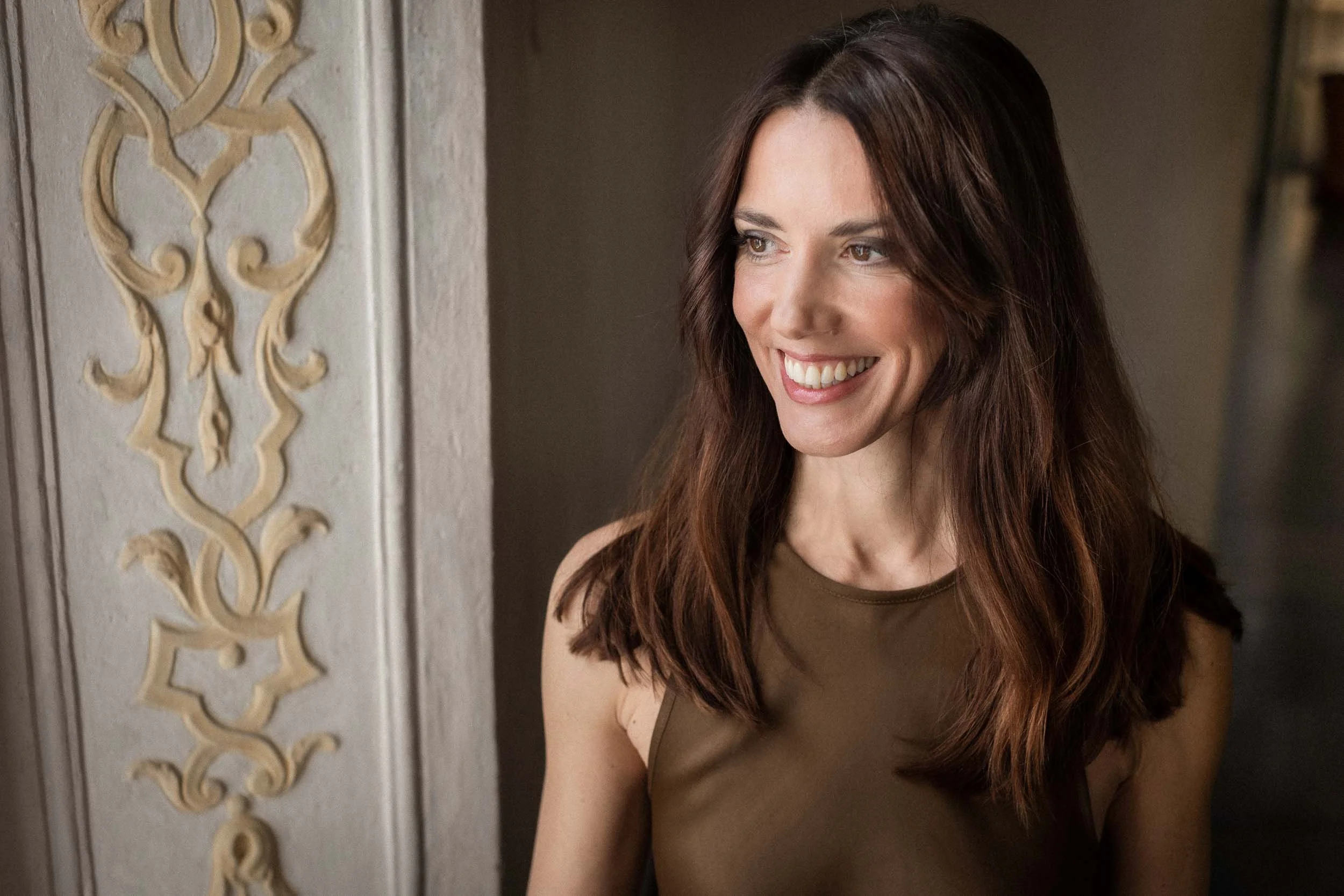 Lächelnde Frau mit braunen, lockigen Haaren, trägt ein braunes Oberteil, steht neben einer verzierte Säule in einem Raum.