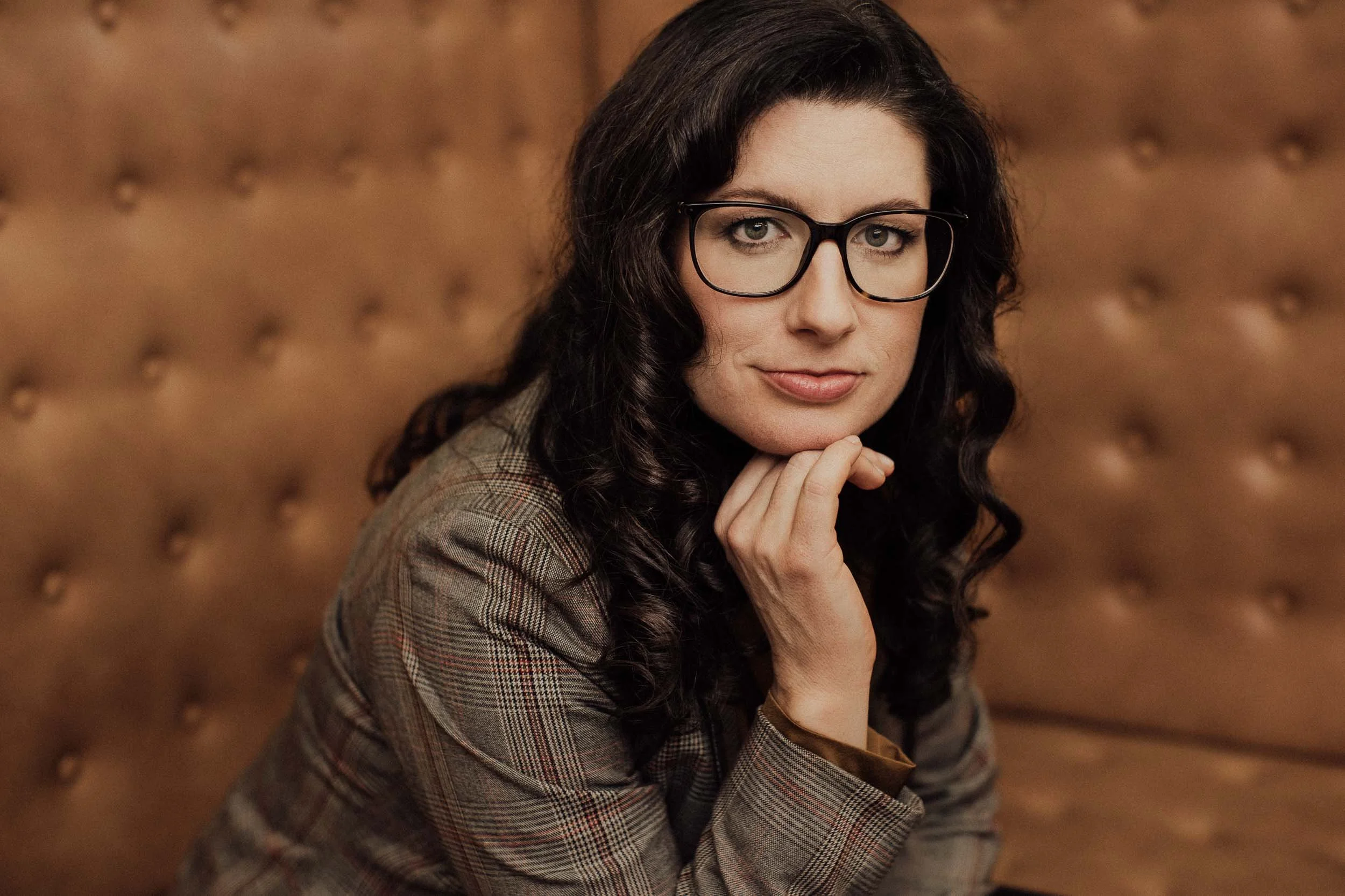 Eine Frau mit dunklen, gewellten Haaren und Brille, die auf einem braunen, gepolsterten Hintergrund sitzt und die Hand an das Kinn legt.