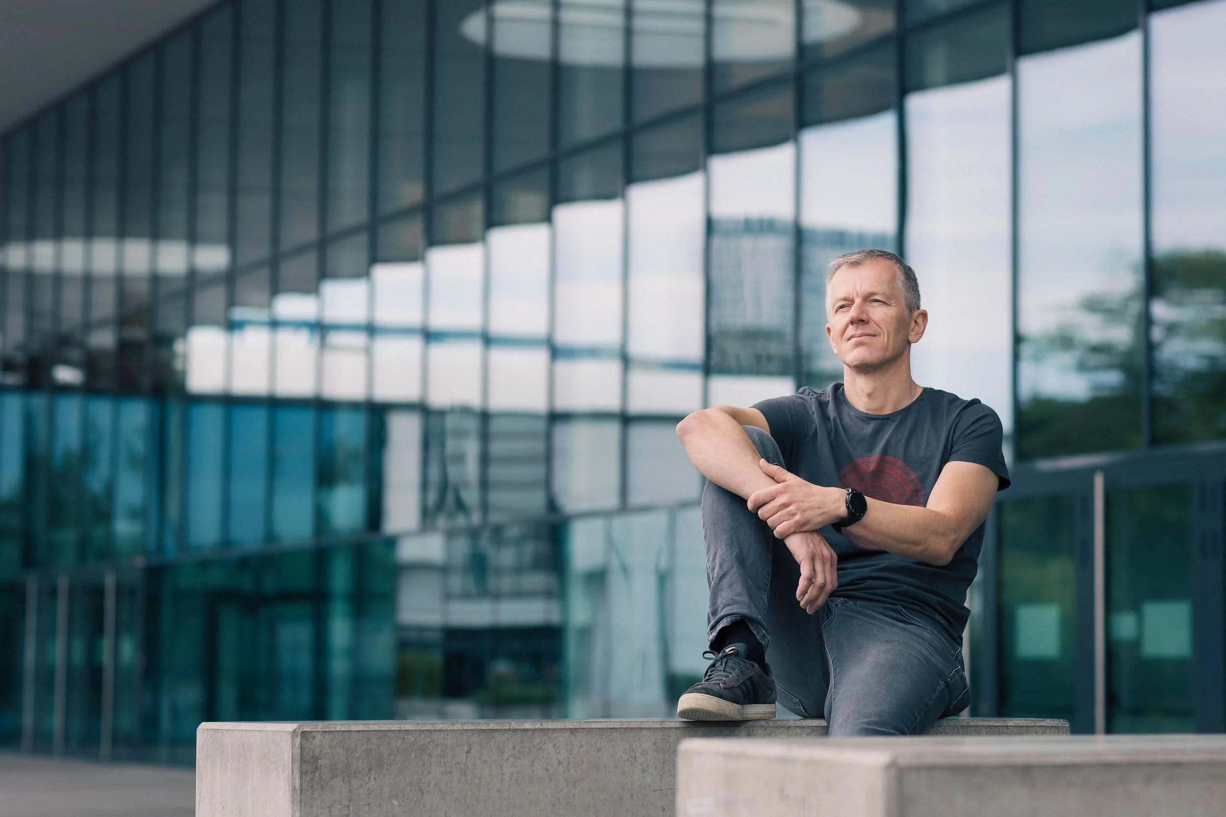 Ein älterer Mann sitzt nachdenklich auf einer Betonbank vor einem modernen Glasgebäude.